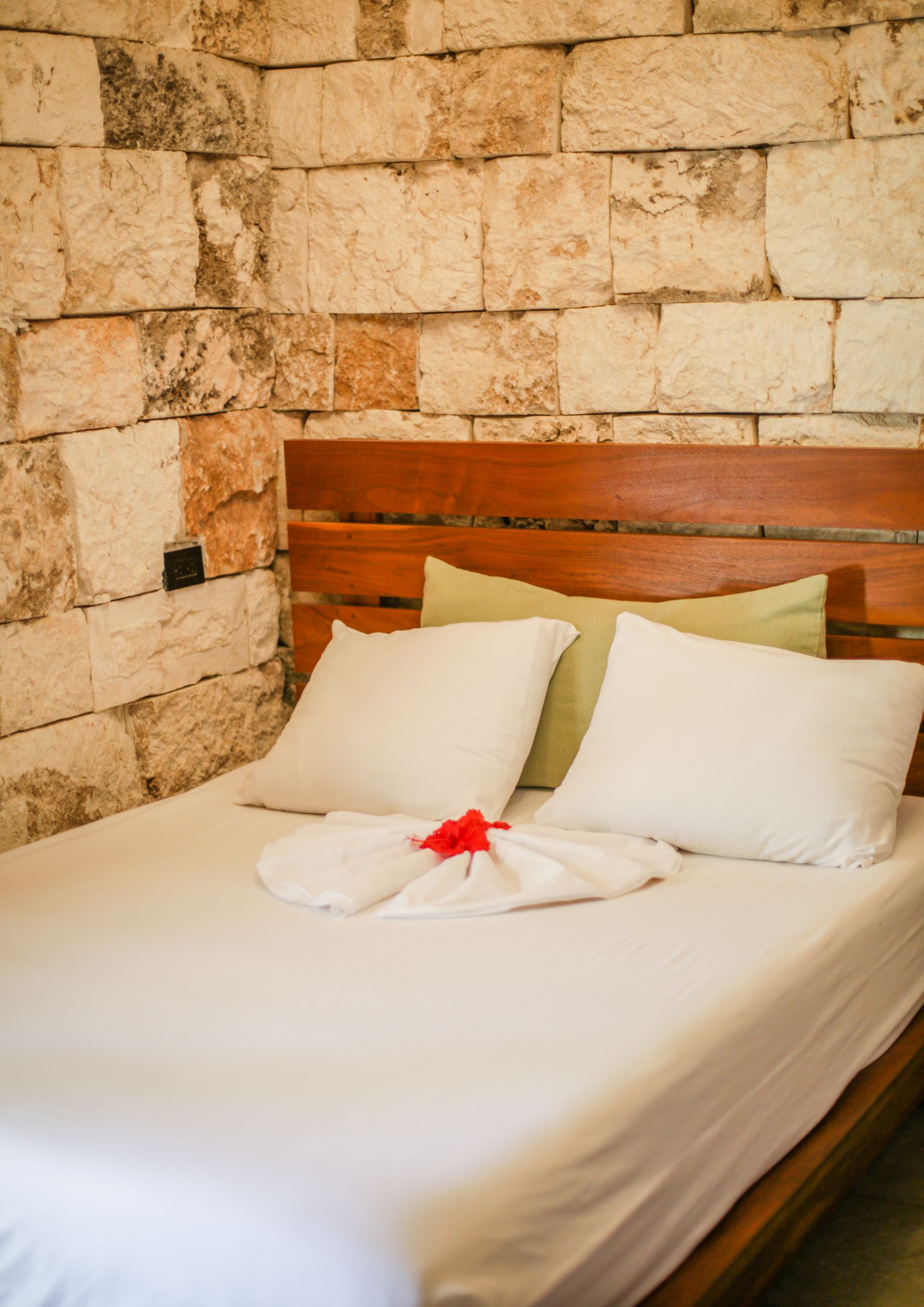 Cama con sábanas blancas, almohadas y toalla decorativa contra una pared de ladrillos de piedra.