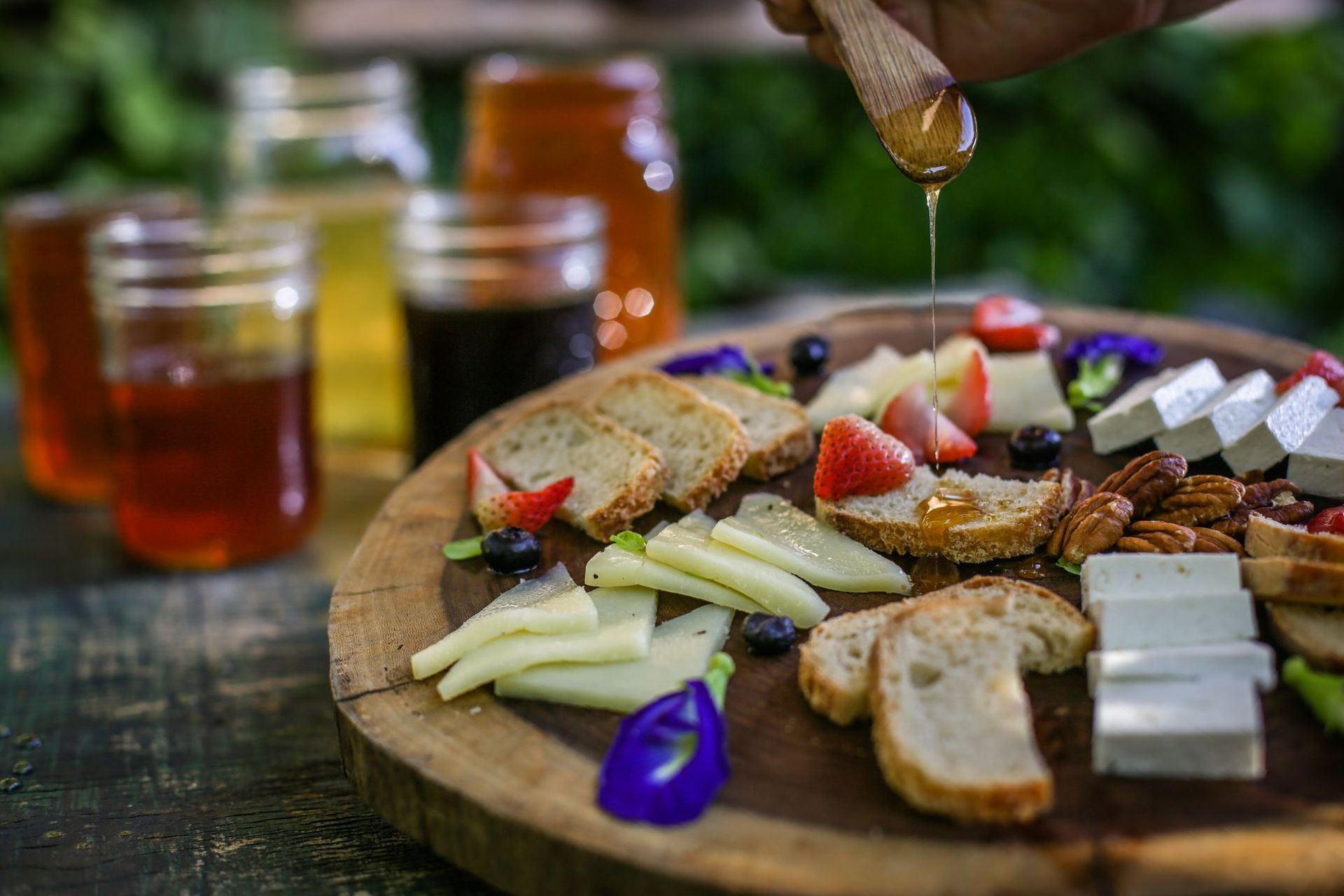 Tabla de embutidos de madera con queso en lonchas, pan, fruta y frutos secos. Se vierte miel con un cucharón de madera. Tarros de miel al fondo.