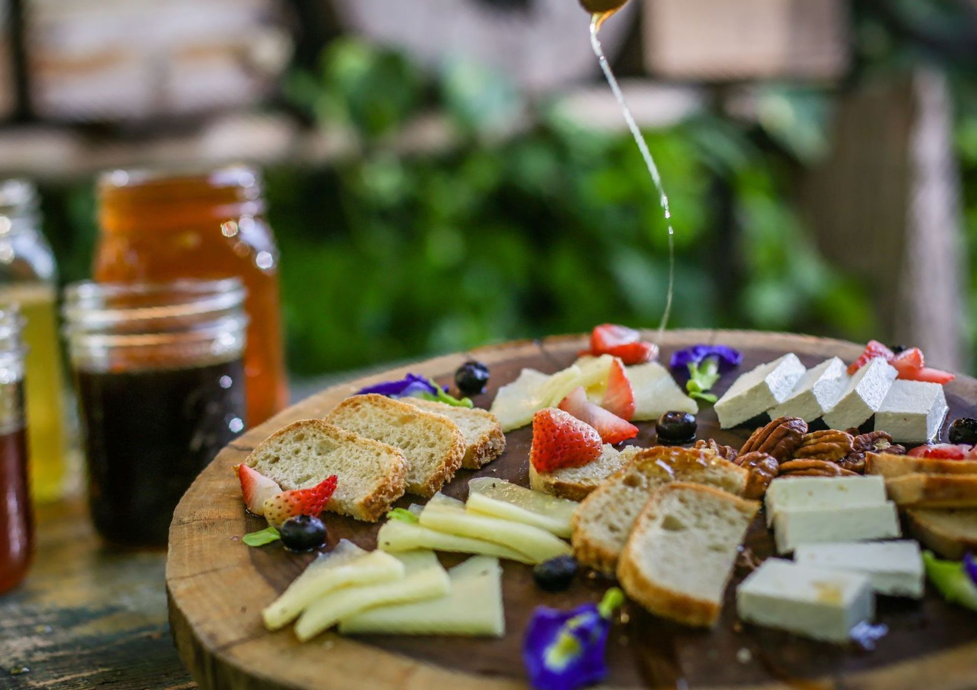 Bandeja de madera con queso, pan, frutas y nueces con miel cayendo al aire libre.