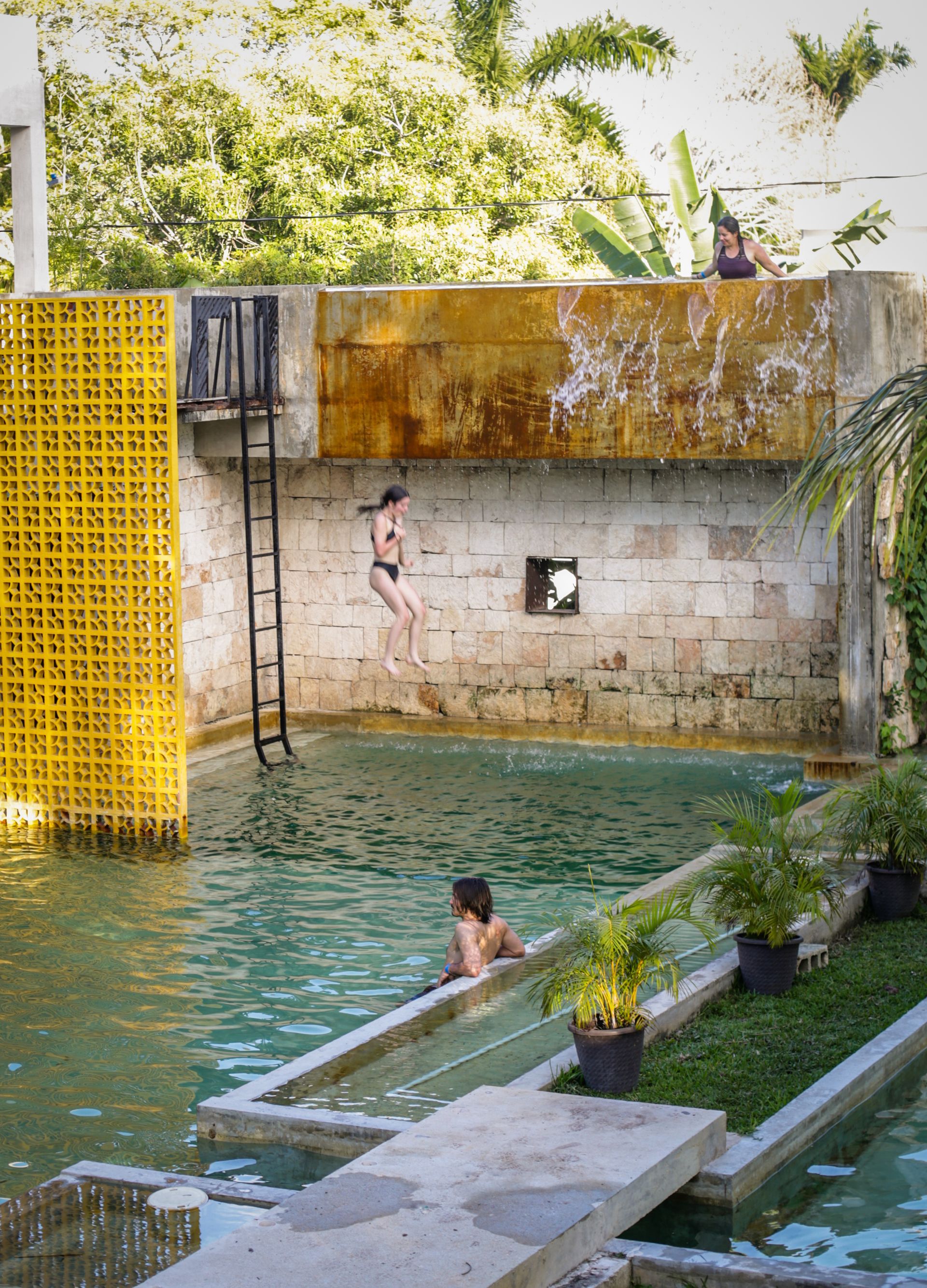 Mujer saltando a una piscina desde una estructura de hormigón. Otras personas en el agua; estructura metálica amarilla a la izquierda; plantas.