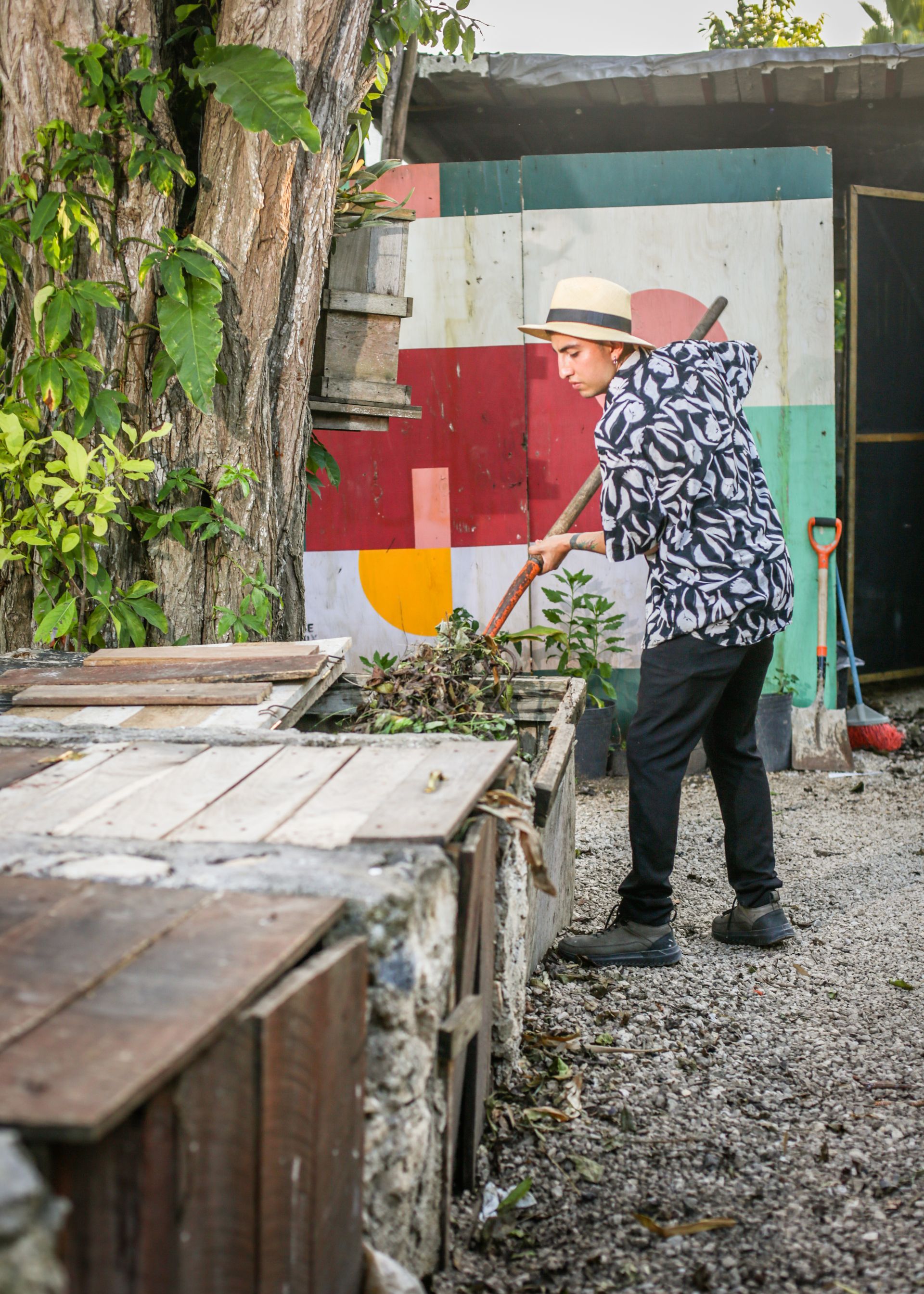 Persona que lleva un sombrero rastrillando hojas en un cantero de jardín con una pared colorida en el fondo.