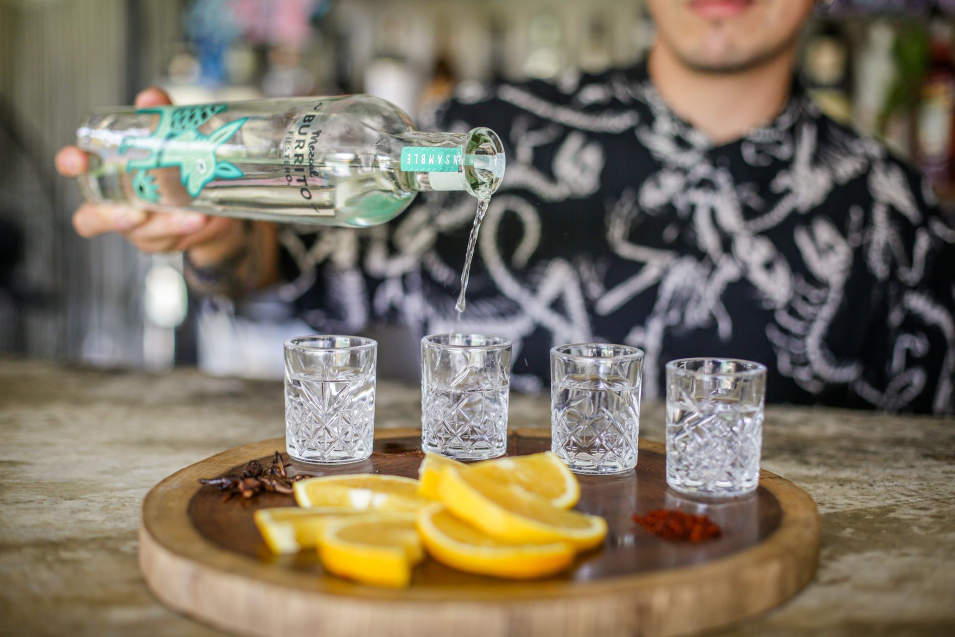 Un camarero sirve líquido de una botella en cuatro vasos de chupito. Rodajas de limón y especias en una bandeja de madera.