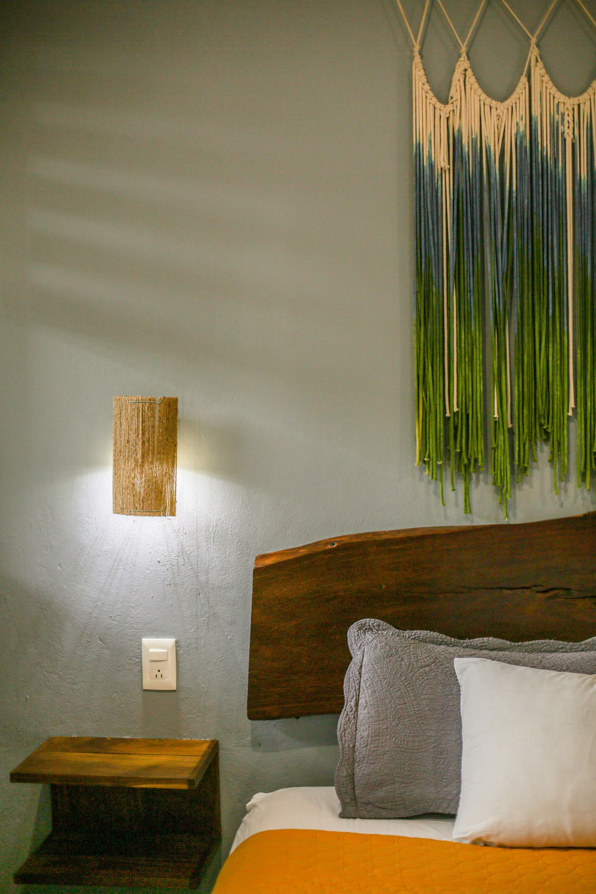 Dormitorio con cabecero de madera, mesita de noche y aplique. Pared verde, manta naranja y tapiz de macramé.