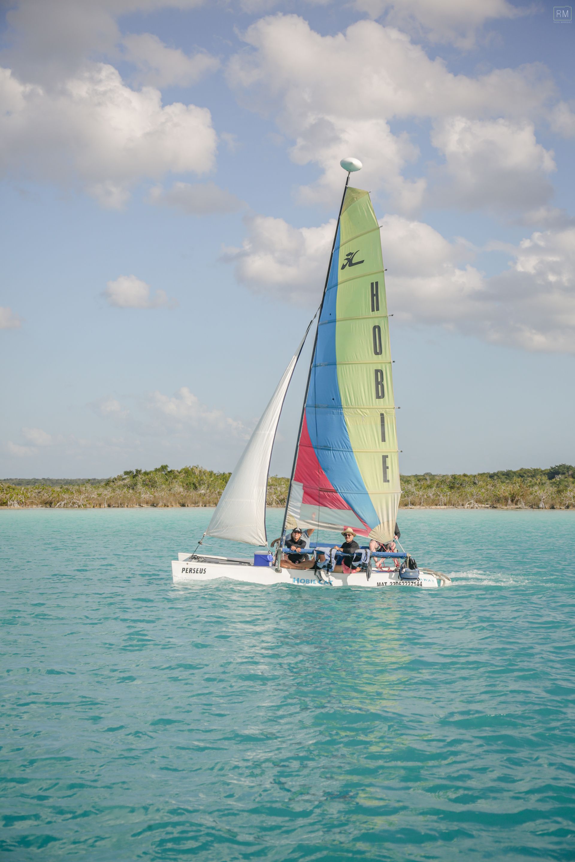 Velero con velas de colores sobre aguas turquesas, con pasajeros a bordo, bajo un cielo azul.