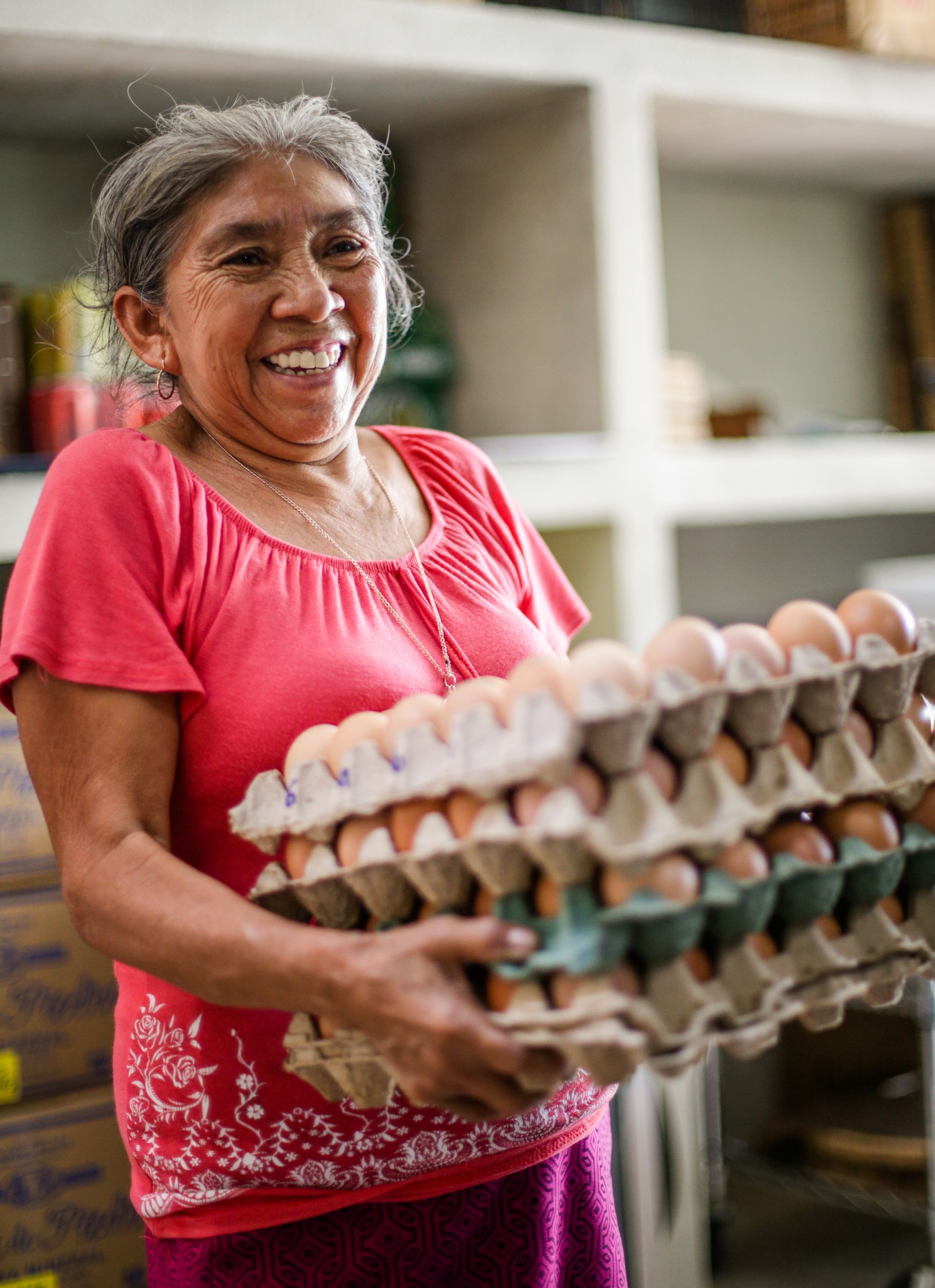 Mujer sonriendo, llevando cartones de huevos apilados en un almacén; camisa roja, estantes abiertos.
