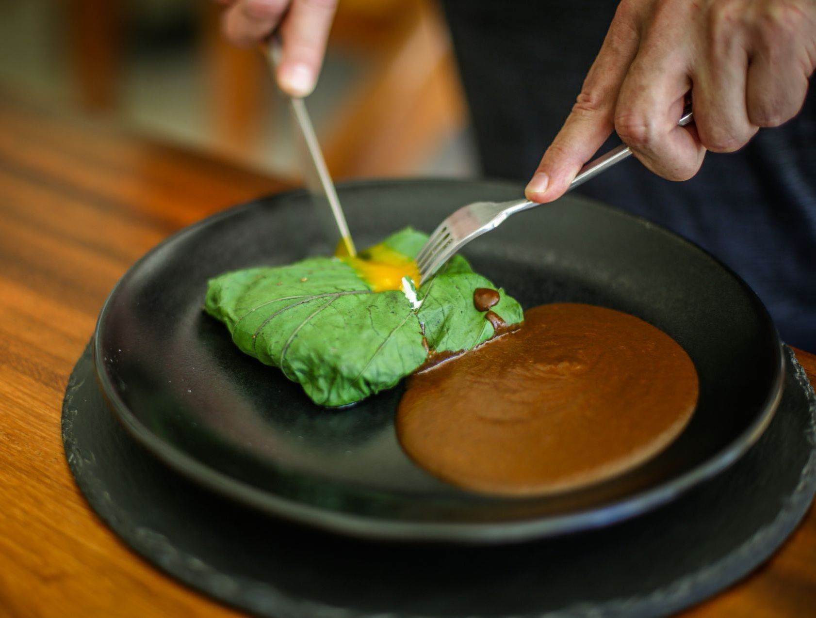 Persona cortando comida envuelta en hojas verdes, con salsa en plato negro.