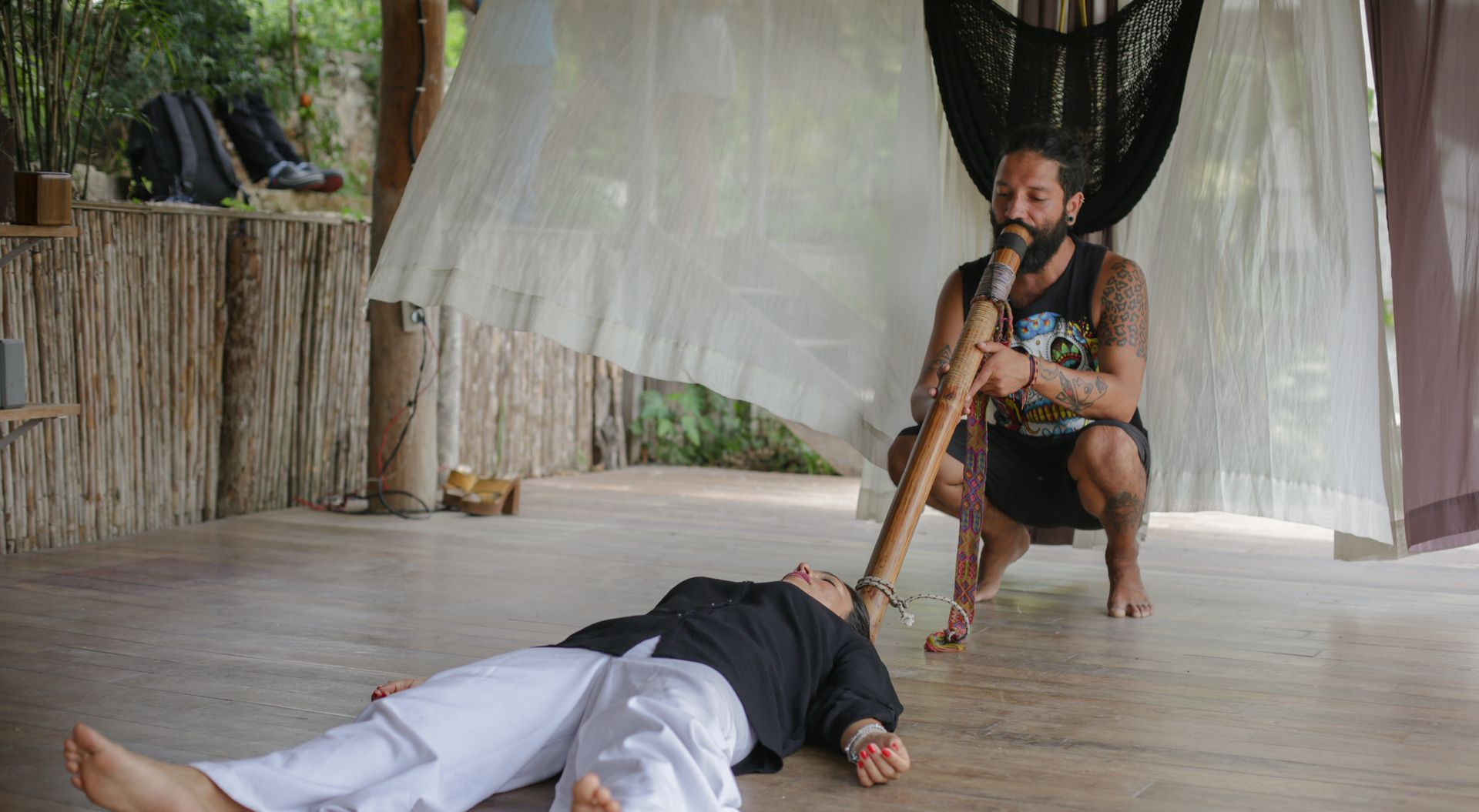 Hombre tocando un didgeridoo junto a una persona tumbada en el suelo, al aire libre. Porche de madera clara.