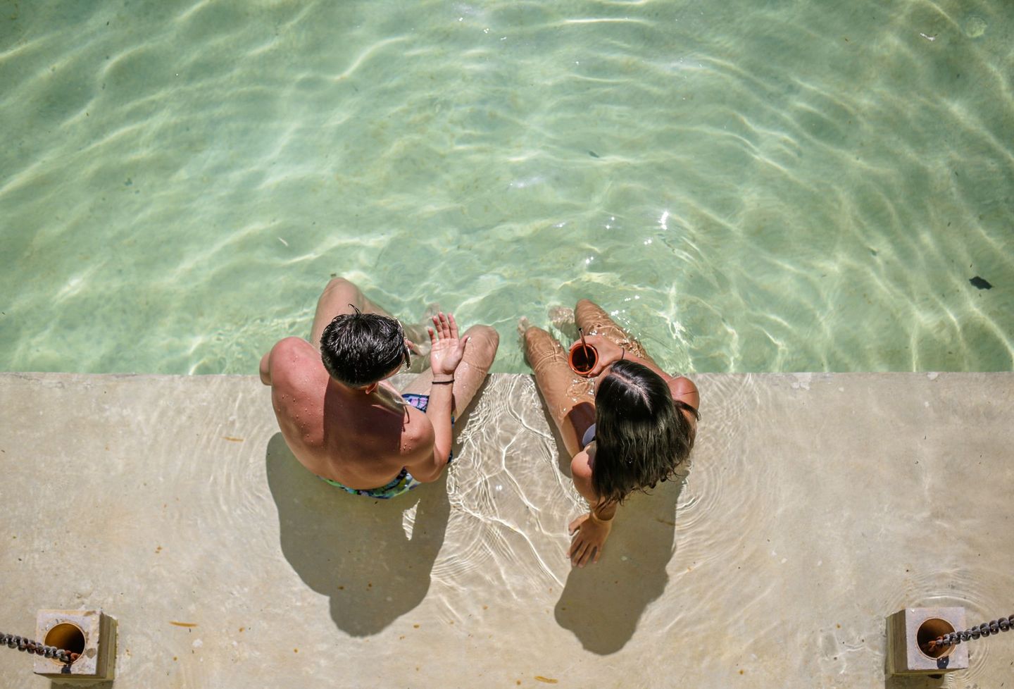 Dos personas sentadas con las piernas juntas en la piscina. La luz del sol brilla en el borde.