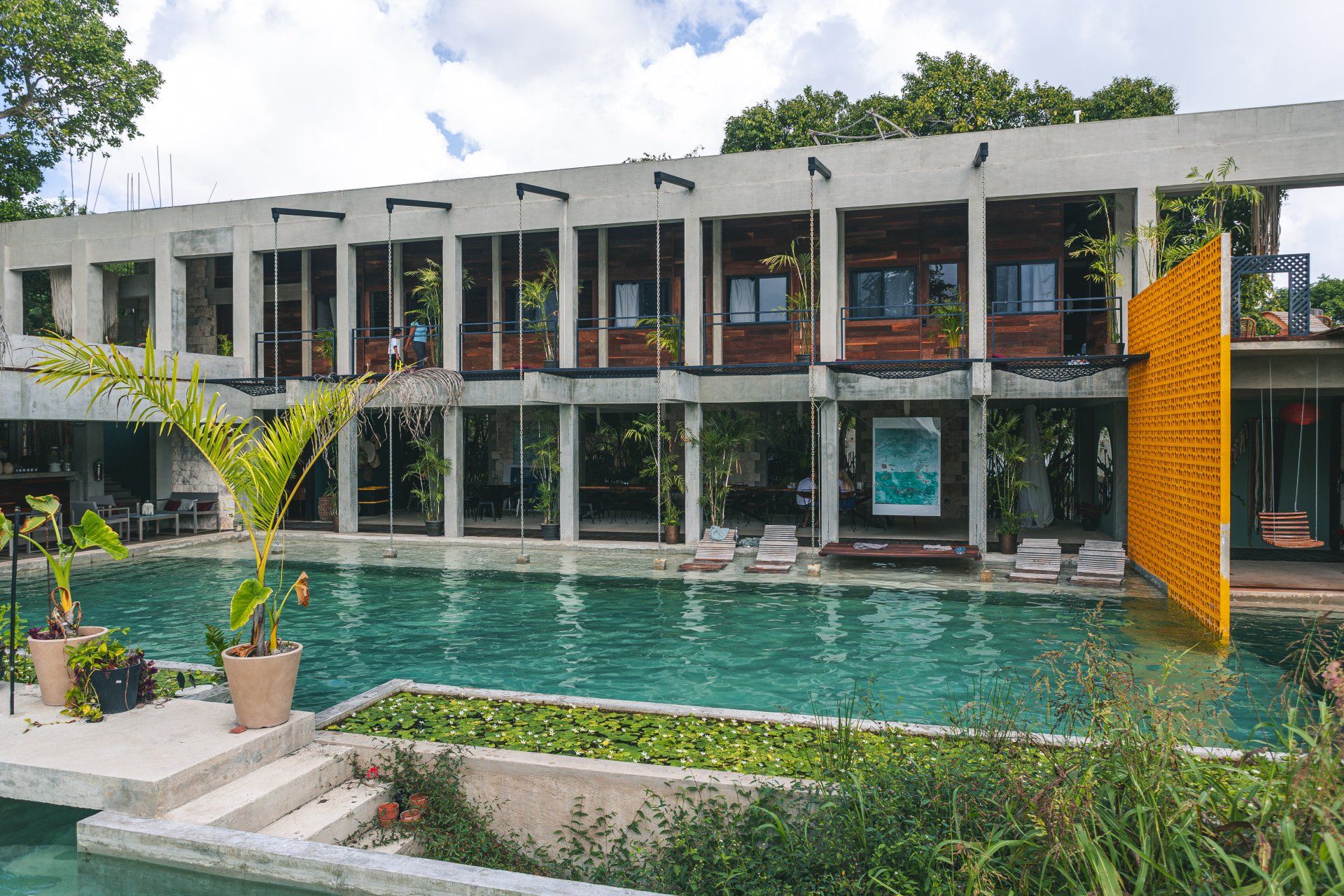 Un edificio moderno con piscina. Hileras de columnas sostienen el segundo piso. Un panel amarillo y árboles enmarcan la escena.