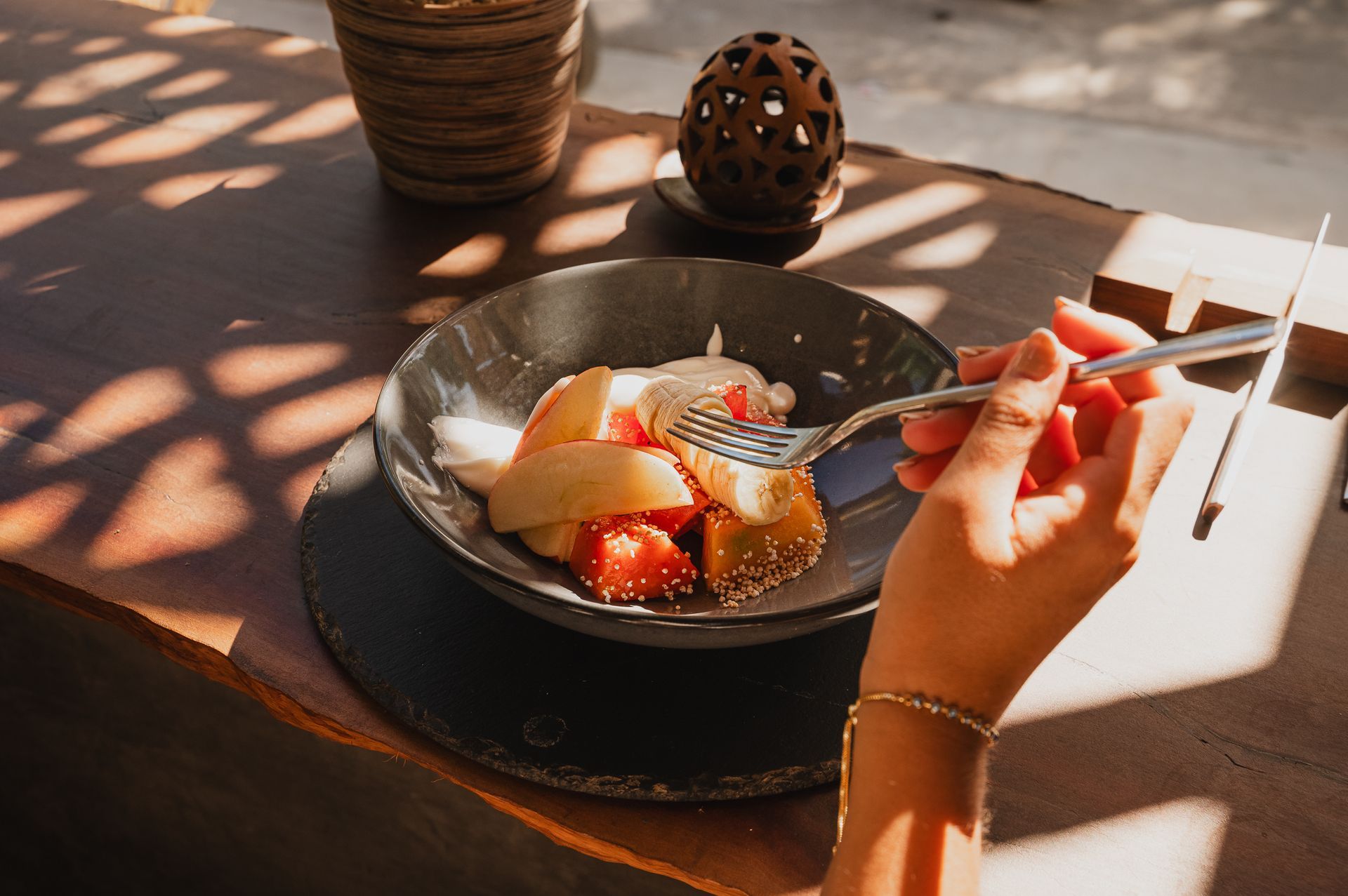 Mano usando un tenedor para comer fruta de un recipiente sobre una mesa de madera, bajo la luz del sol.