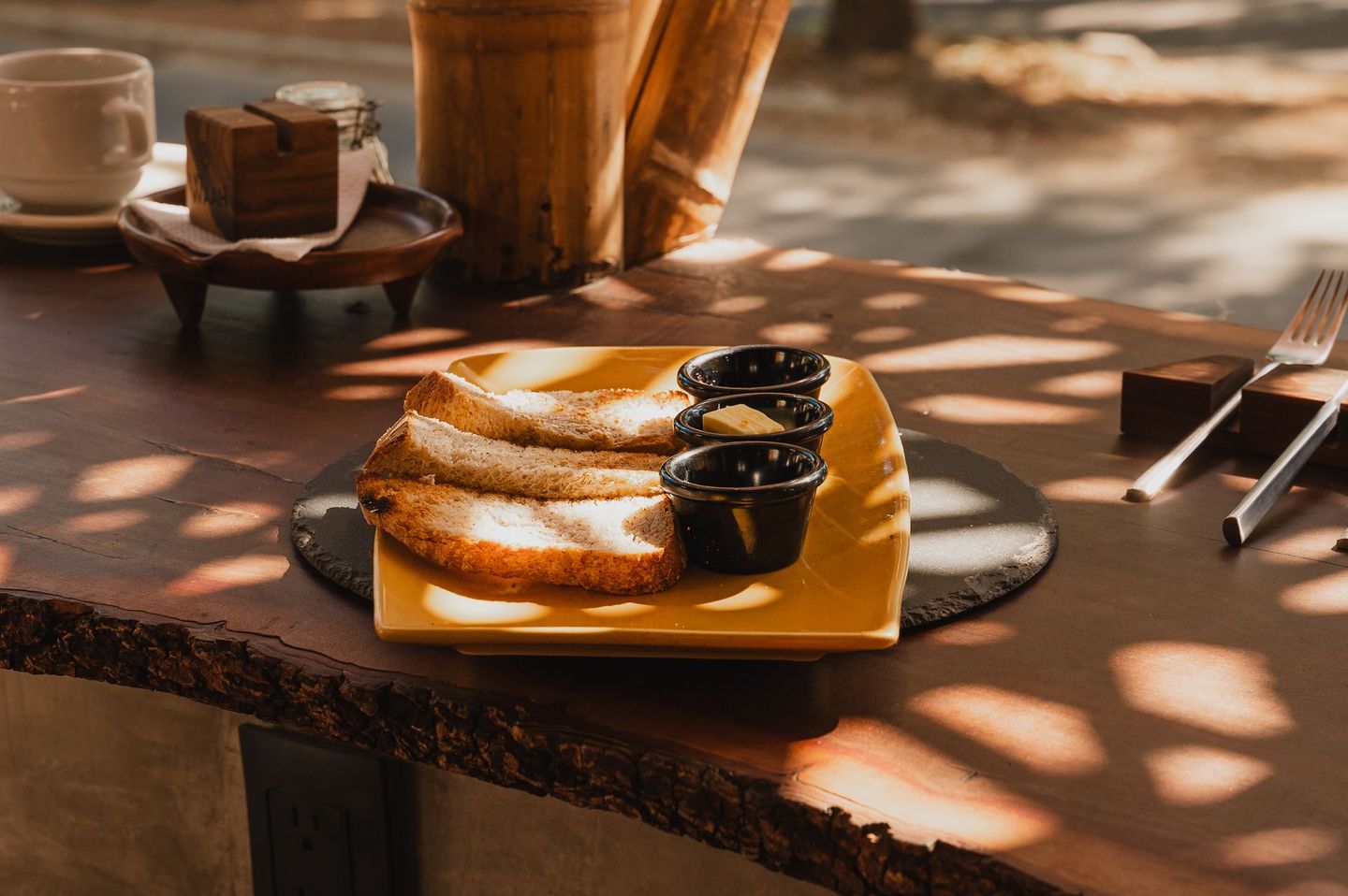 Panqueques en un plato amarillo con salsas aparte, una taza y un tenedor sobre una mesa de madera con luz moteada.