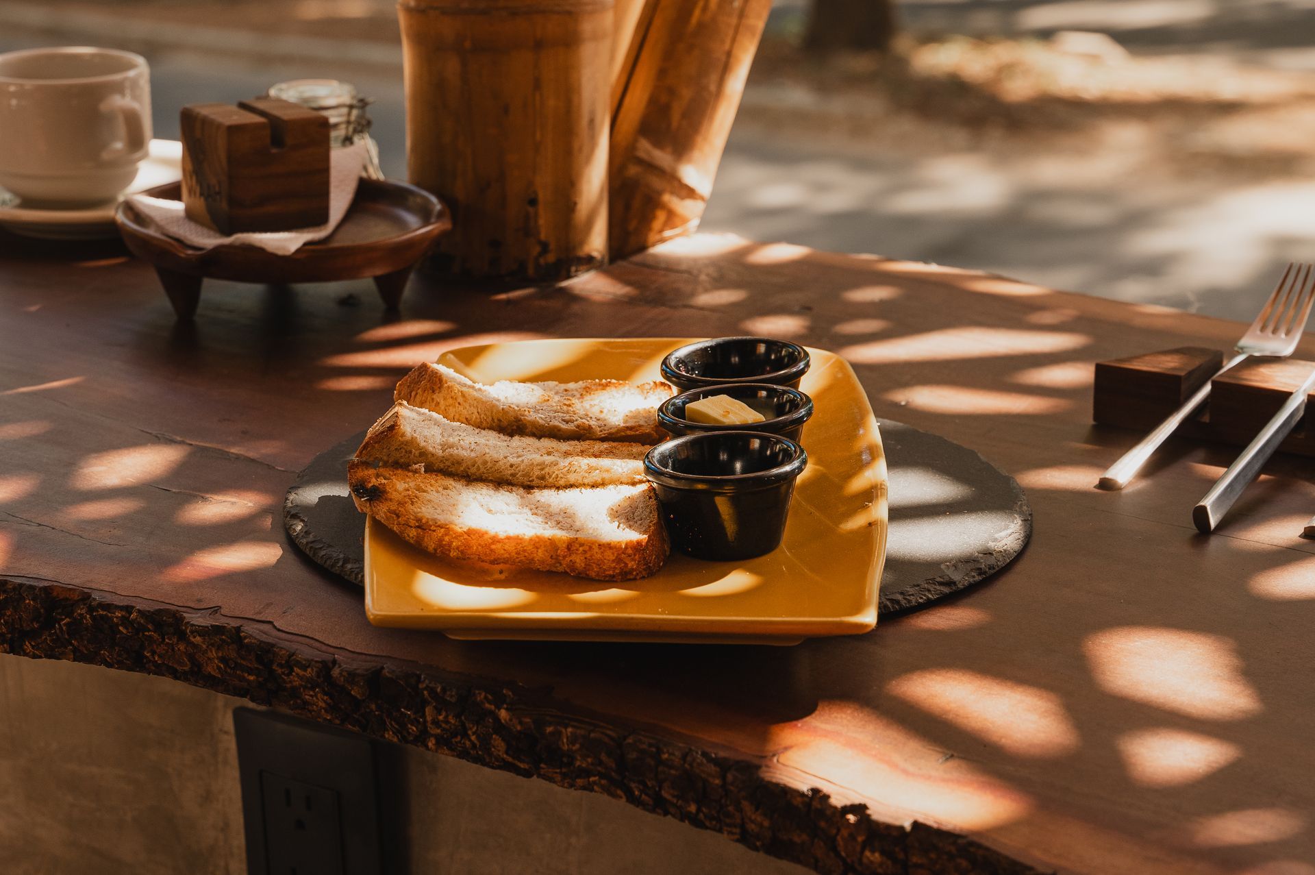 Panqueques en un plato amarillo con salsas aparte, una taza y un tenedor sobre una mesa de madera con luz moteada.