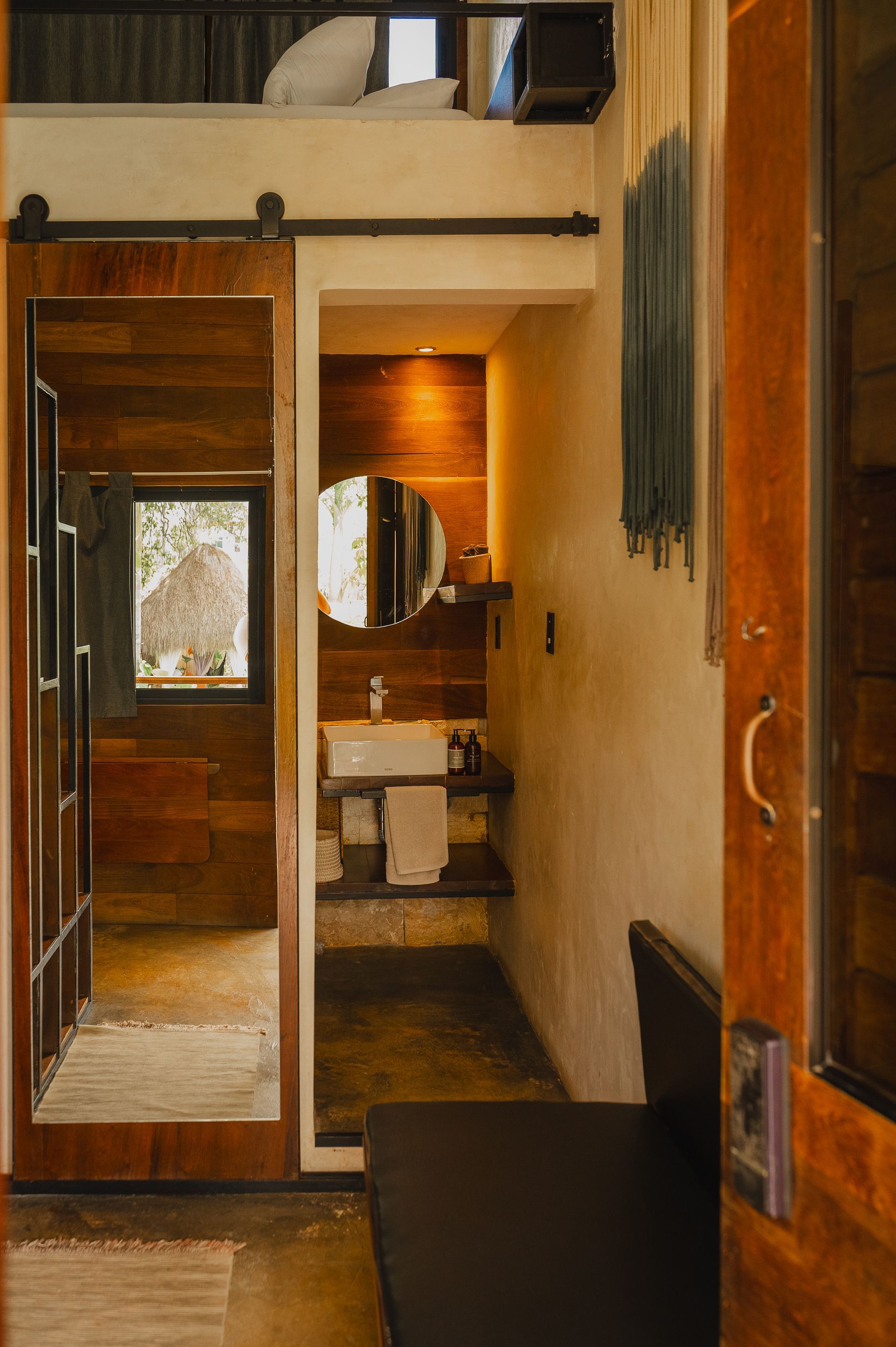 Interior acogedor con cama alta, baño con lavabo y puerta corredera de granero. Madera natural y tonos neutros.