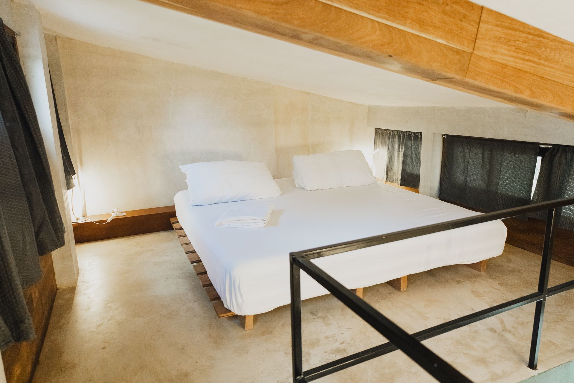 Dormitorio con cama queen en un altillo. La habitación tiene paredes en tonos neutros y vigas de madera. Una barandilla negra se encuentra en primer plano.
