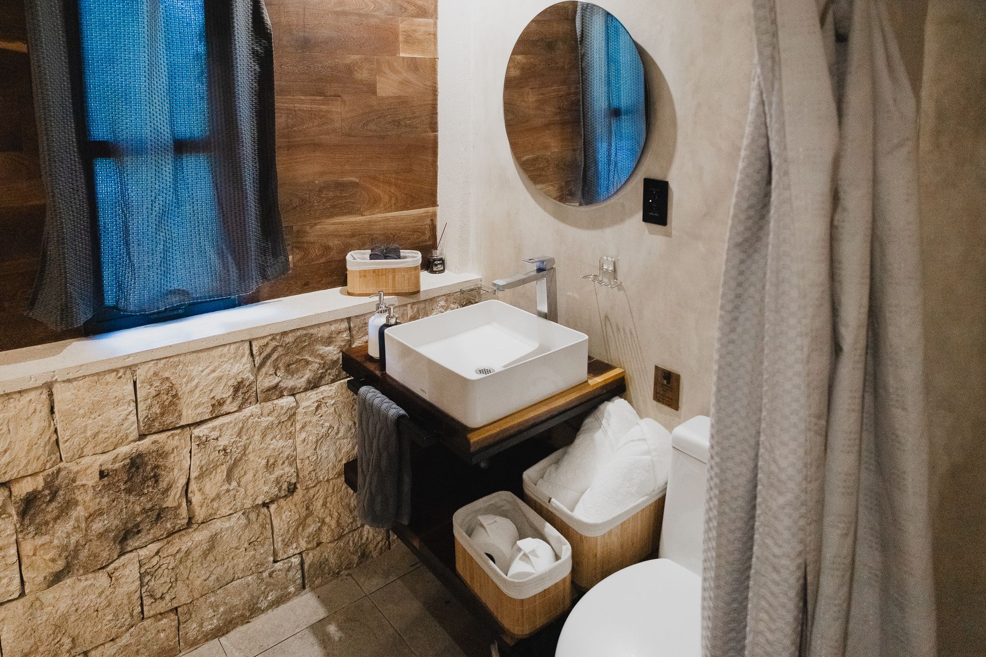 Baño con pared de piedra, lavabo, espejo e inodoro. Los tonos suaves y neutros crean un ambiente de spa.