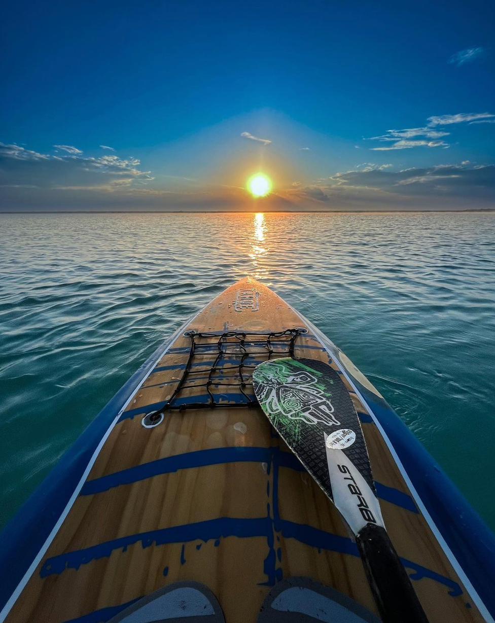 Paddleboarder en el agua hacia el atardecer. Remo en el marco, tabla de madera, agua y cielo azules, y sol dorado.