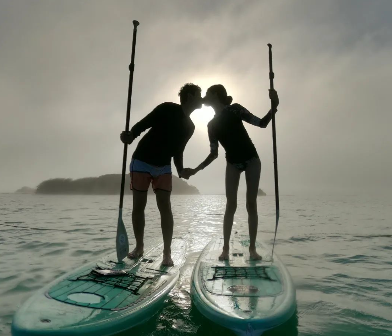 Pareja besándose en tablas de paddle surf frente al sol, tomados de la mano, recortados en el agua.