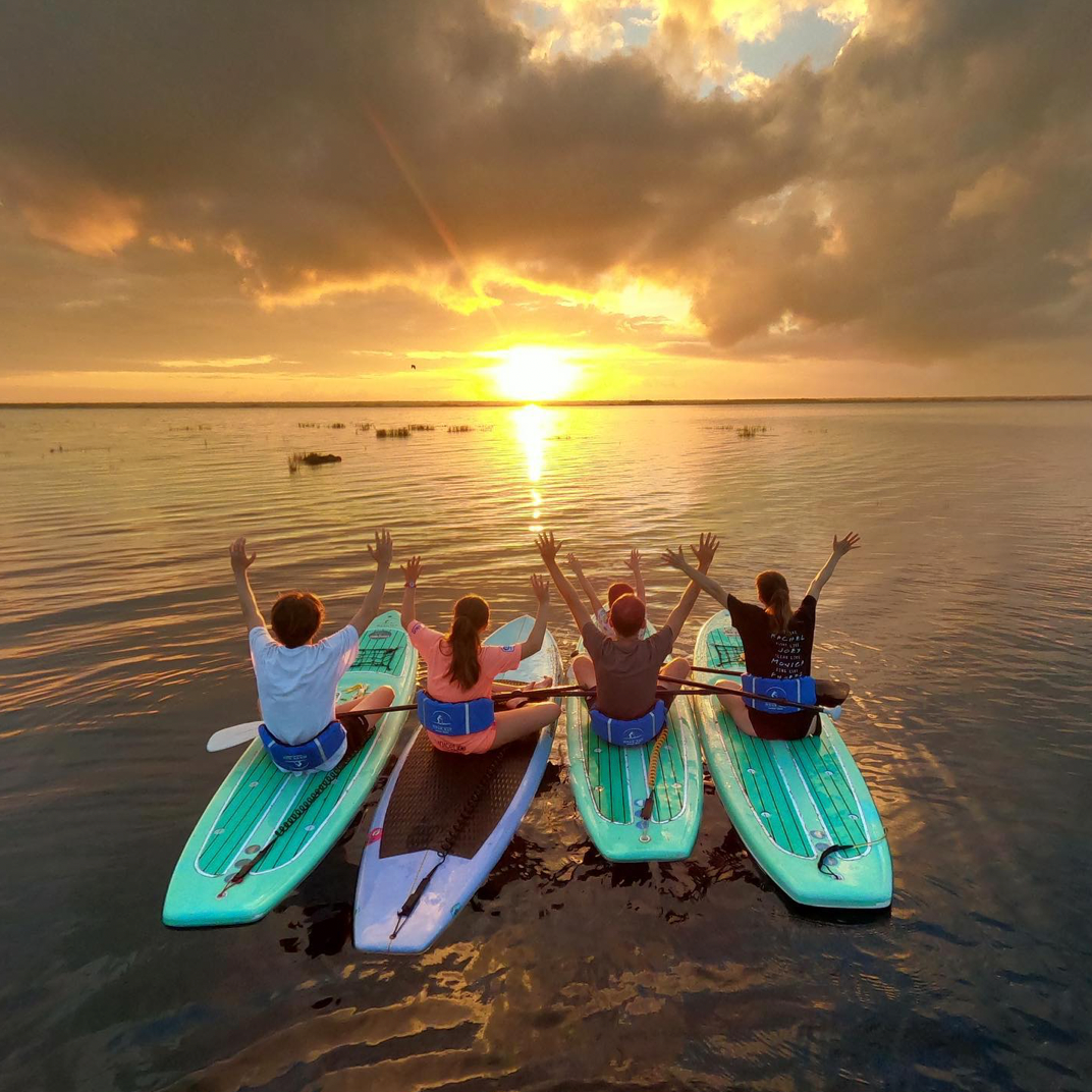 Cuatro personas en tablas de paddle surf, con los brazos en alto, contemplando la puesta de sol sobre aguas tranquilas.