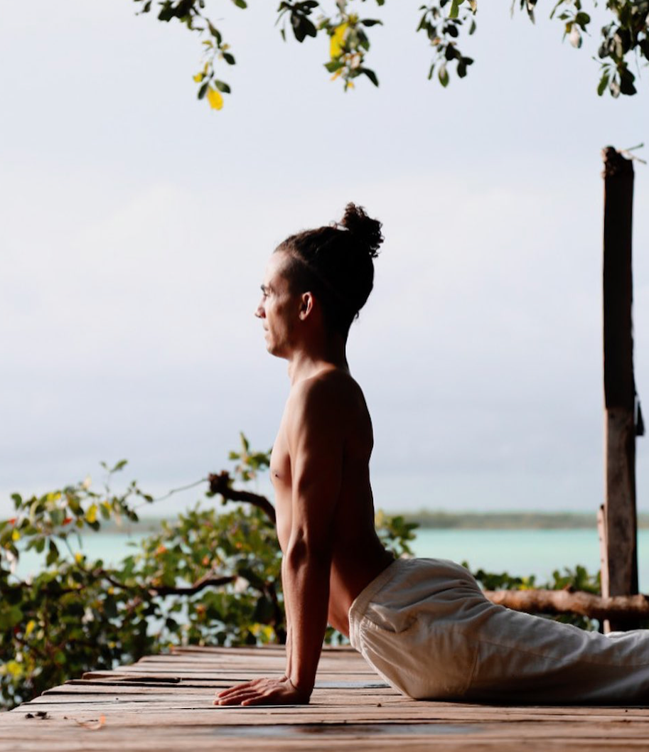 Hombre en pose de yoga al aire libre en una plataforma de madera cerca del agua.