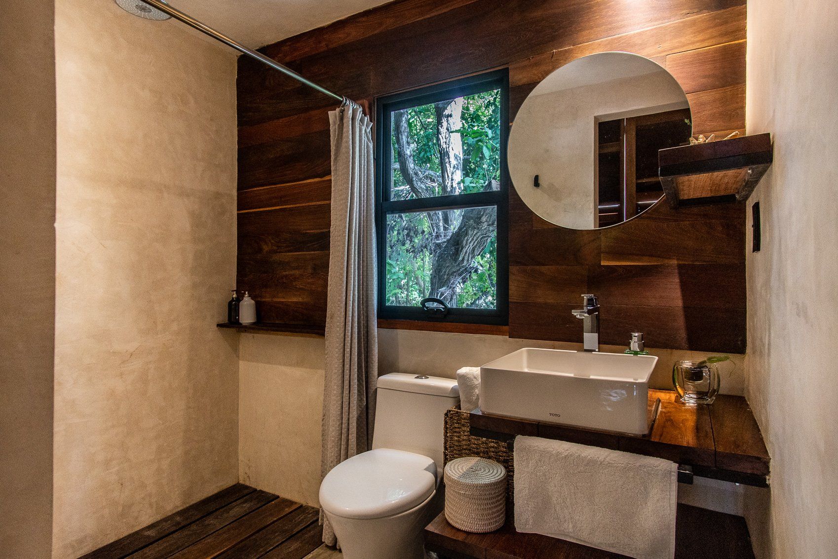 Baño rústico con paneles de madera, lavabo blanco, inodoro, espejo redondo y una ventana con vista a los árboles.