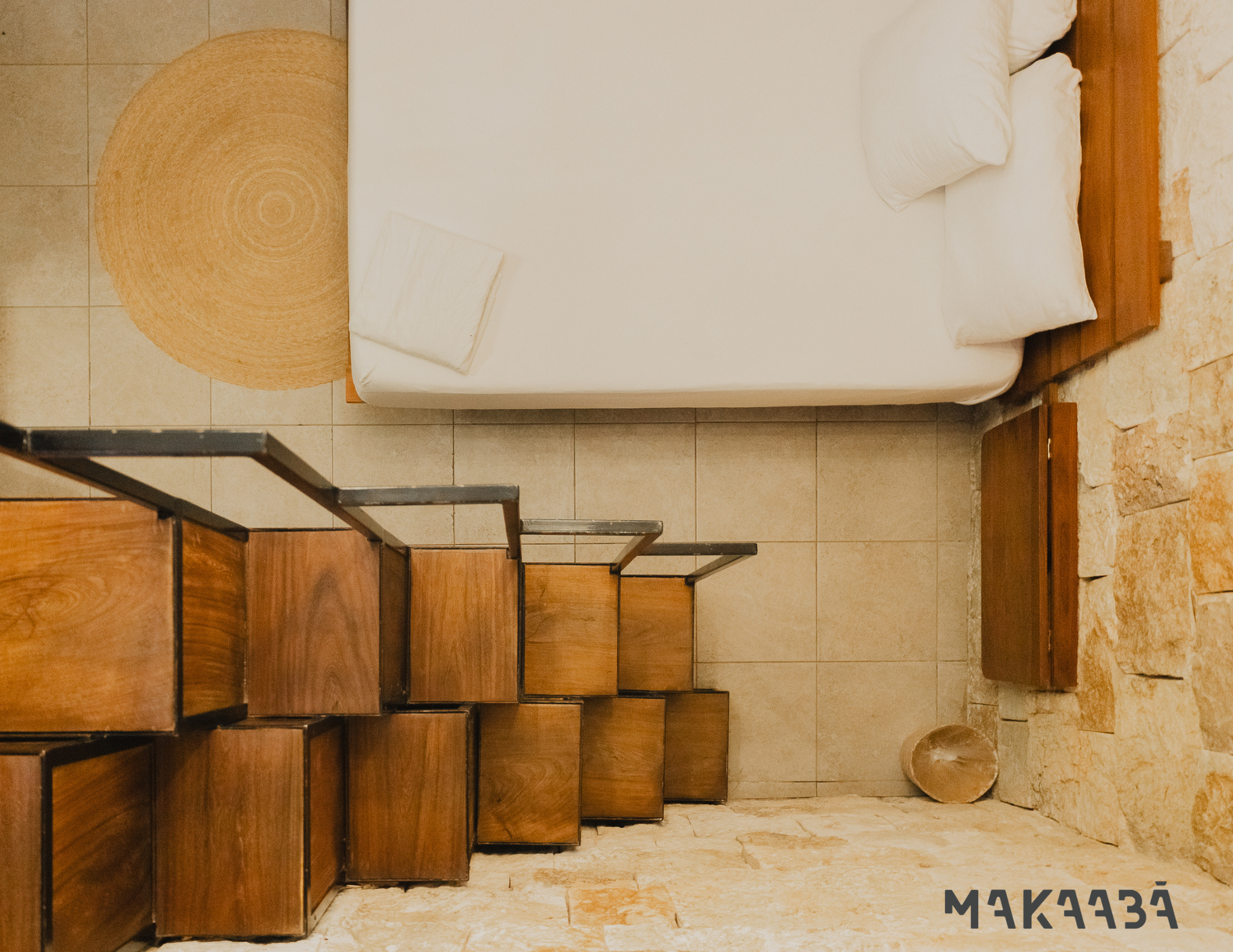 Dormitorio con escalera de madera, cama y pared de piedra. Alfombra de yute en el suelo.