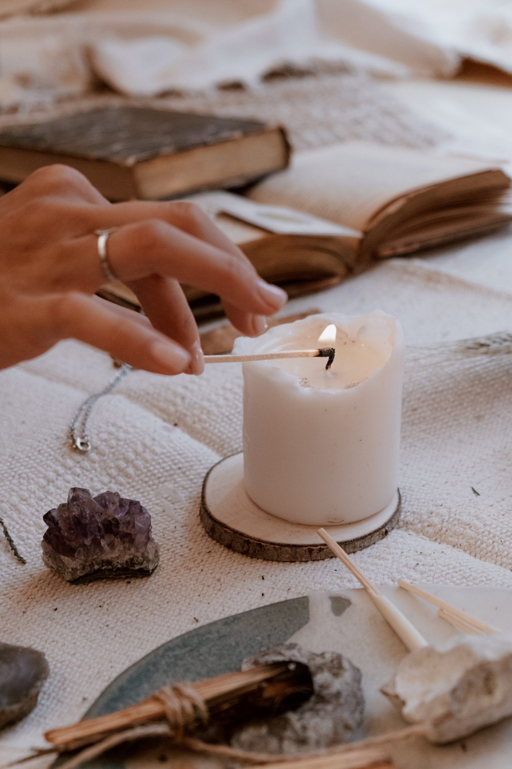 Mano encendiendo una vela blanca sobre un posavasos de madera. Amatista, libro abierto y otros objetos sobre tela blanca texturizada.