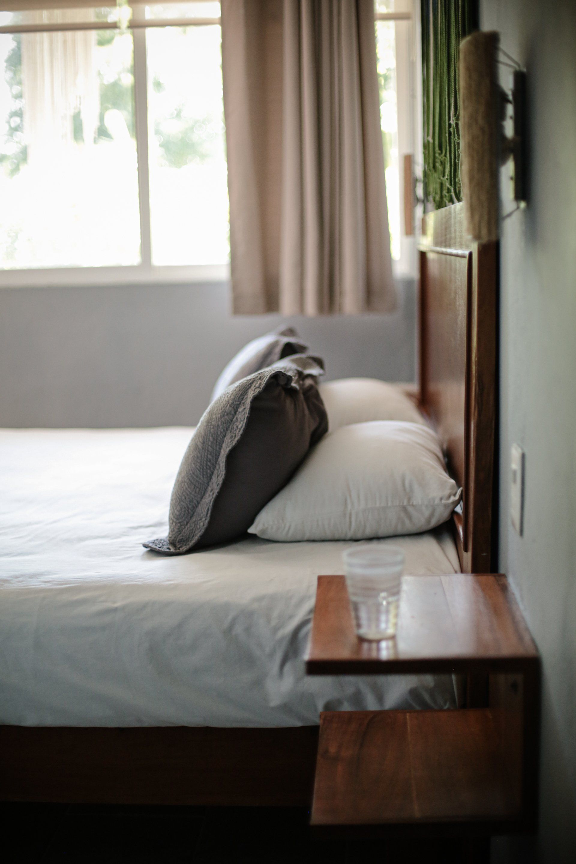 Cama con ropa de cama blanca y gris, dos almohadas, cabecero y mesita de noche de madera, vaso de agua.