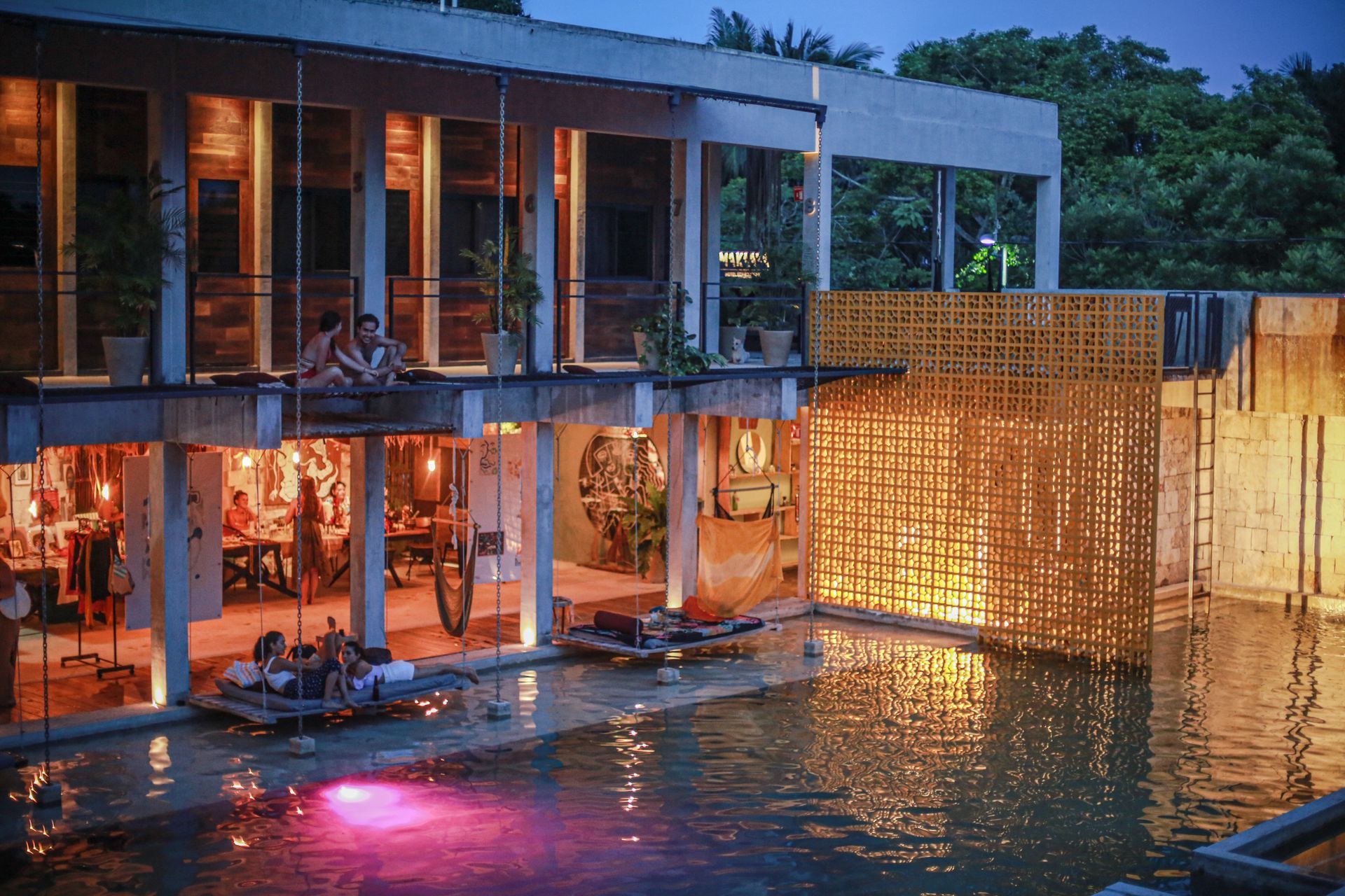 Escena nocturna: gente en un restaurante con piscina. El edificio cuenta con terrazas, estructuras de madera iluminadas y agua con luces reflectantes.