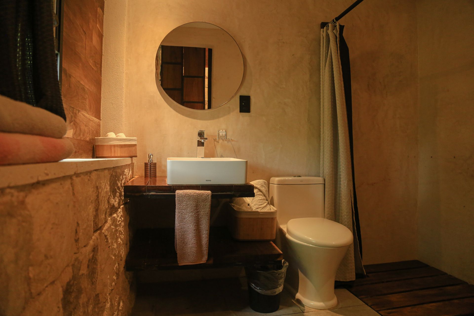 Baño con pared de piedra, lavabo de madera, espejo redondo e inodoro.