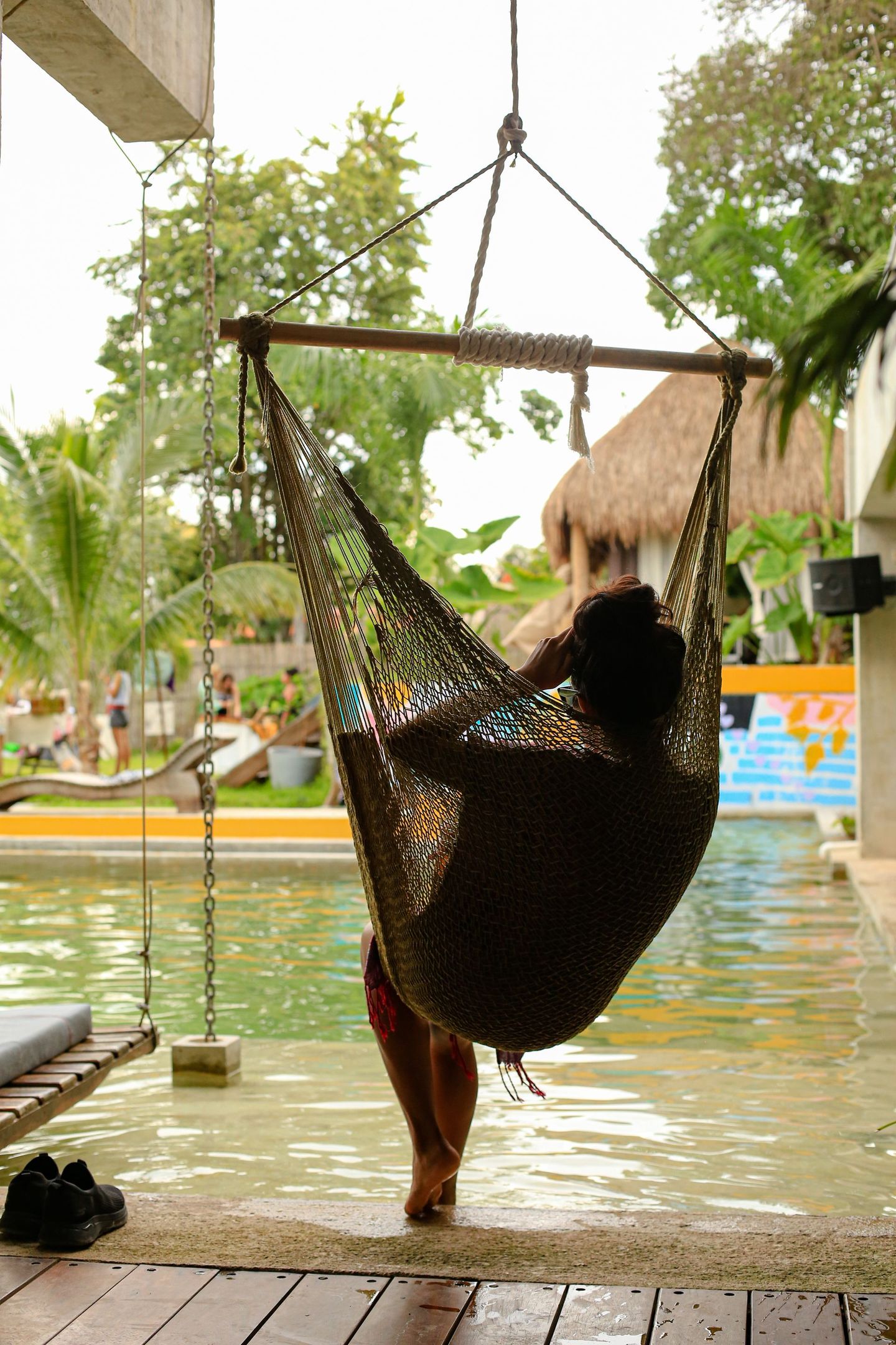 Persona descansando en una hamaca cerca de una piscina. La hamaca es marrón, el agua de la piscina es verde y el entorno es exuberante.