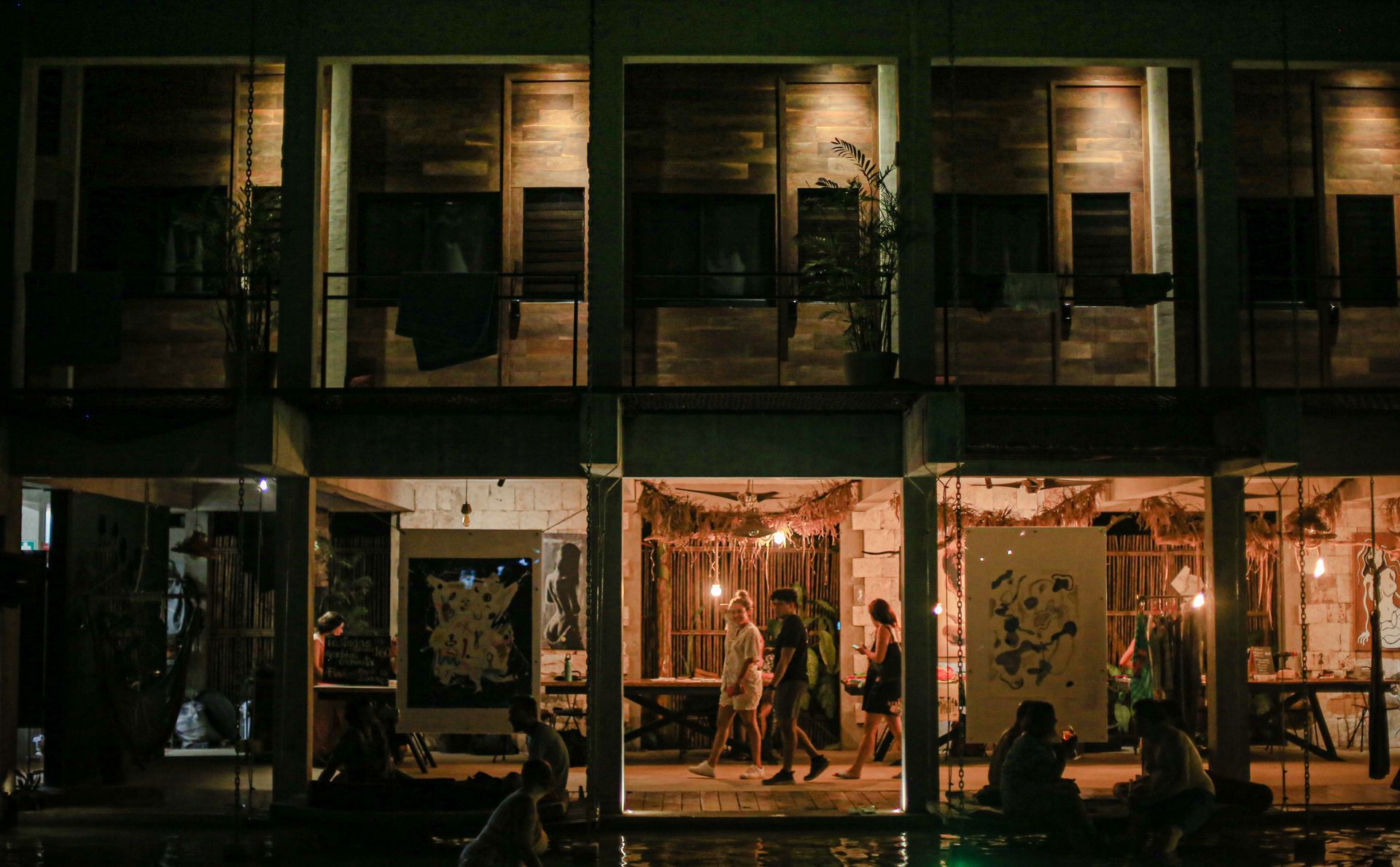 Vista exterior de un edificio iluminado de varios pisos por la noche, con gente caminando en las habitaciones y una piscina al frente.