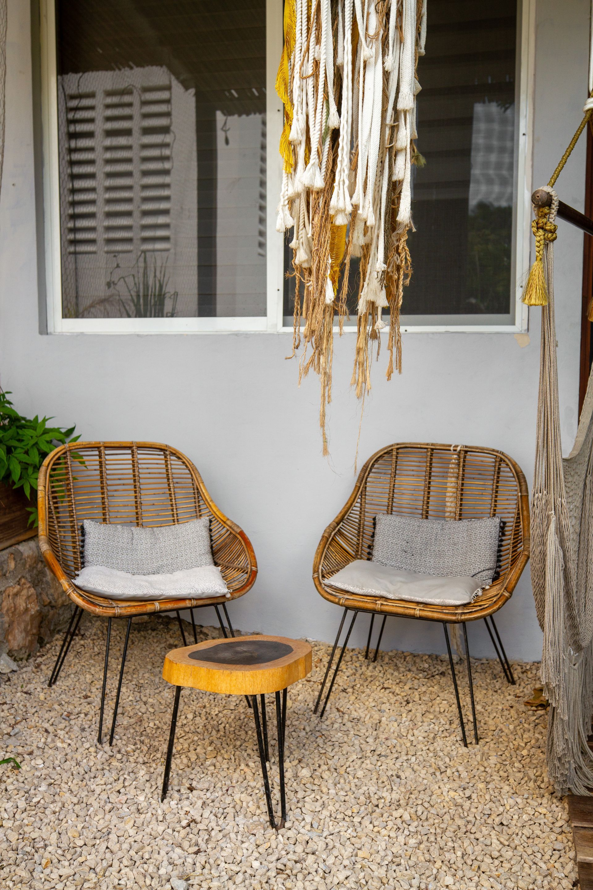Dos sillas de mimbre y una mesa pequeña en un patio de guijarros, con un colgante de macramé y una ventana al fondo.