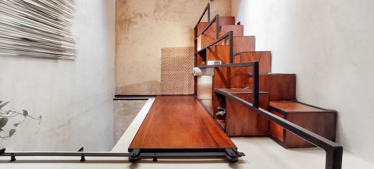 Escalera de madera con espacio de almacenamiento y barandilla integrados. Estructura de madera marrón y metal negro sobre paredes blancas con textura.