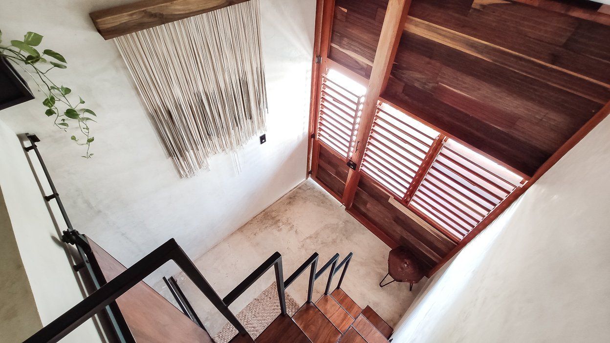 Escalera con escalones y barandilla de madera que conduce al nivel superior. Tapiz de macramé y techo de madera visibles.
