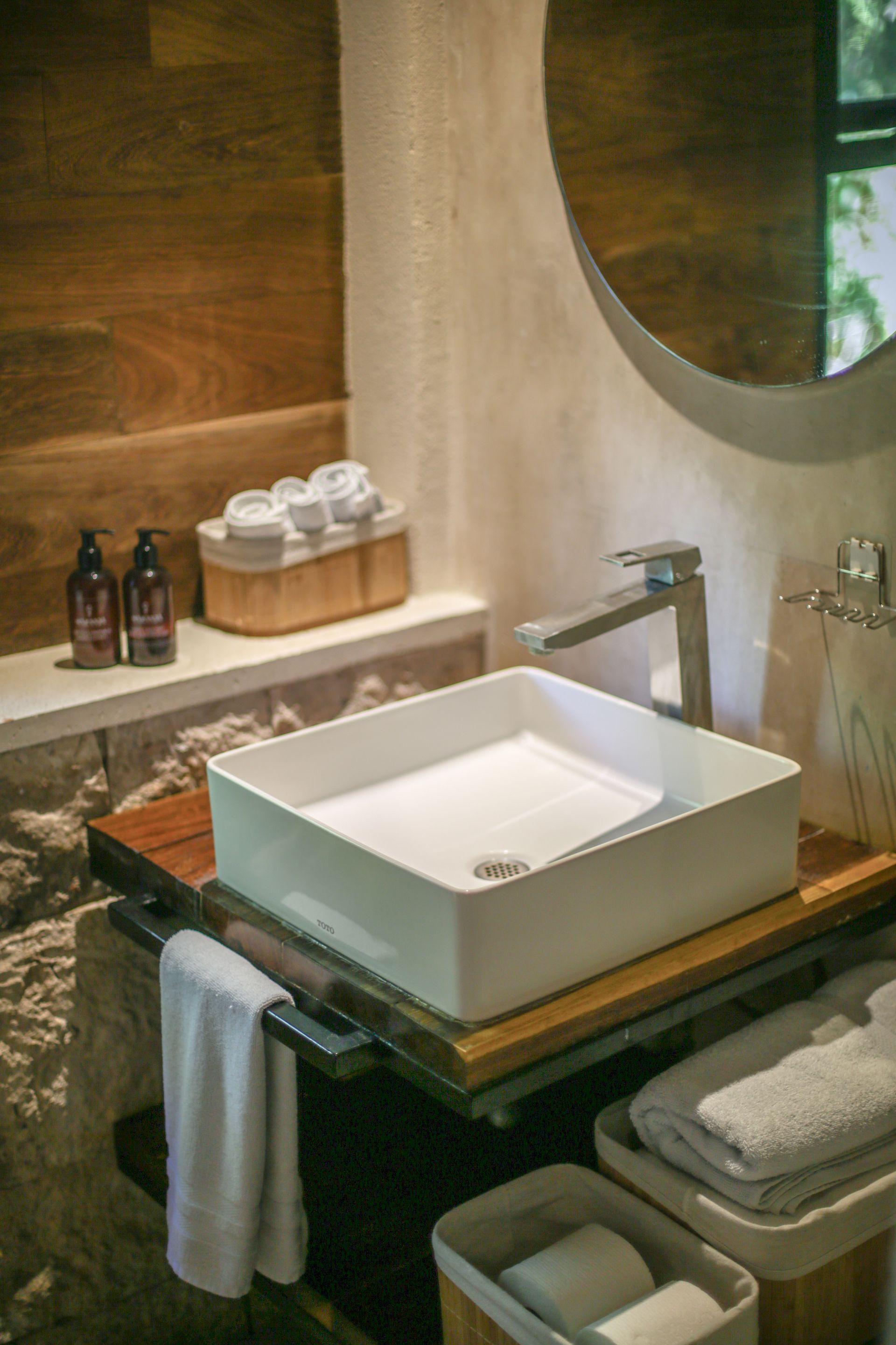 Baño con lavabo cuadrado blanco sobre encimera de madera, toallas y espejo redondo.