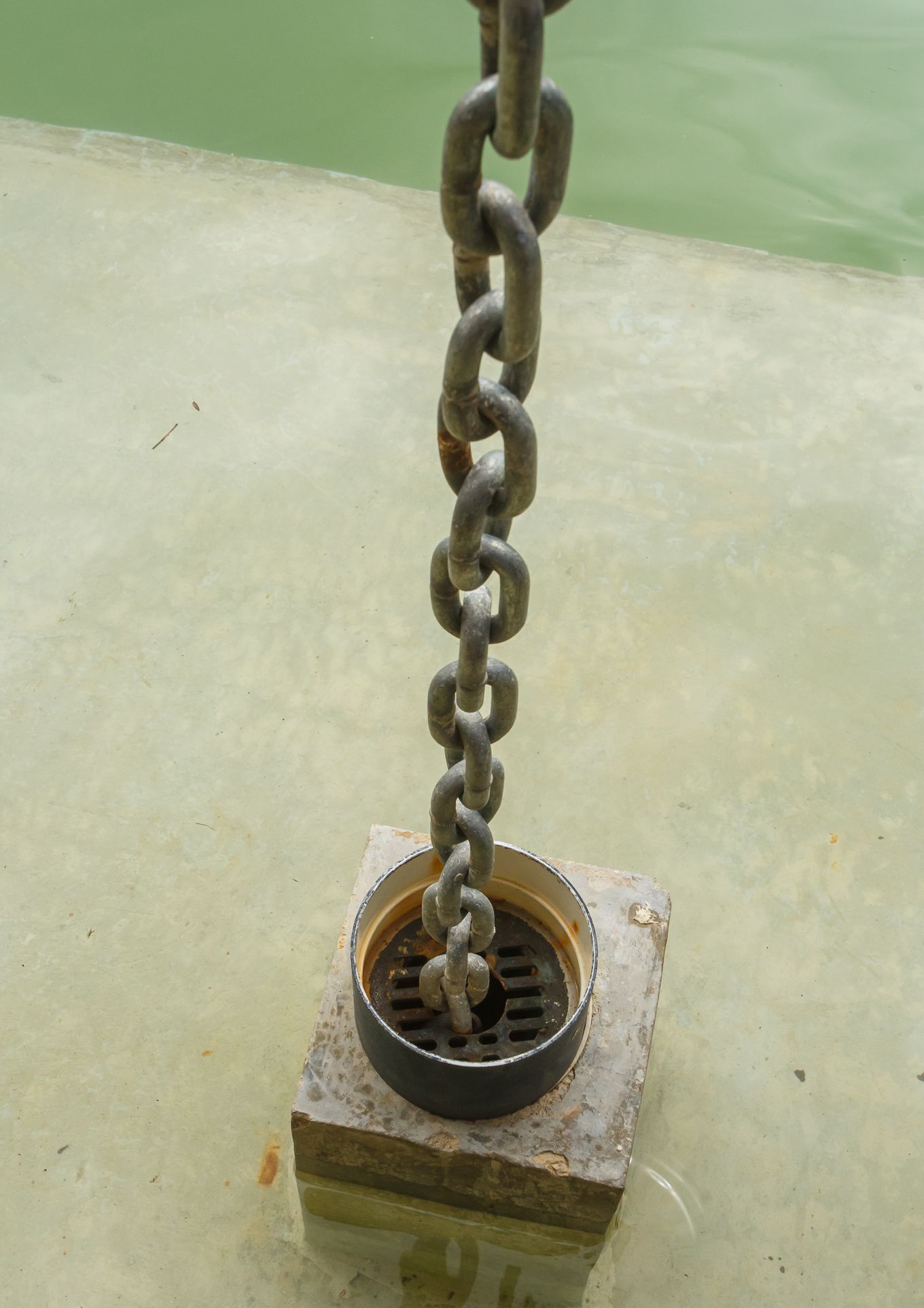 Una cadena de metal cuelga de un desagüe situado sobre un bloque de hormigón, el fondo es una superficie verde claro.