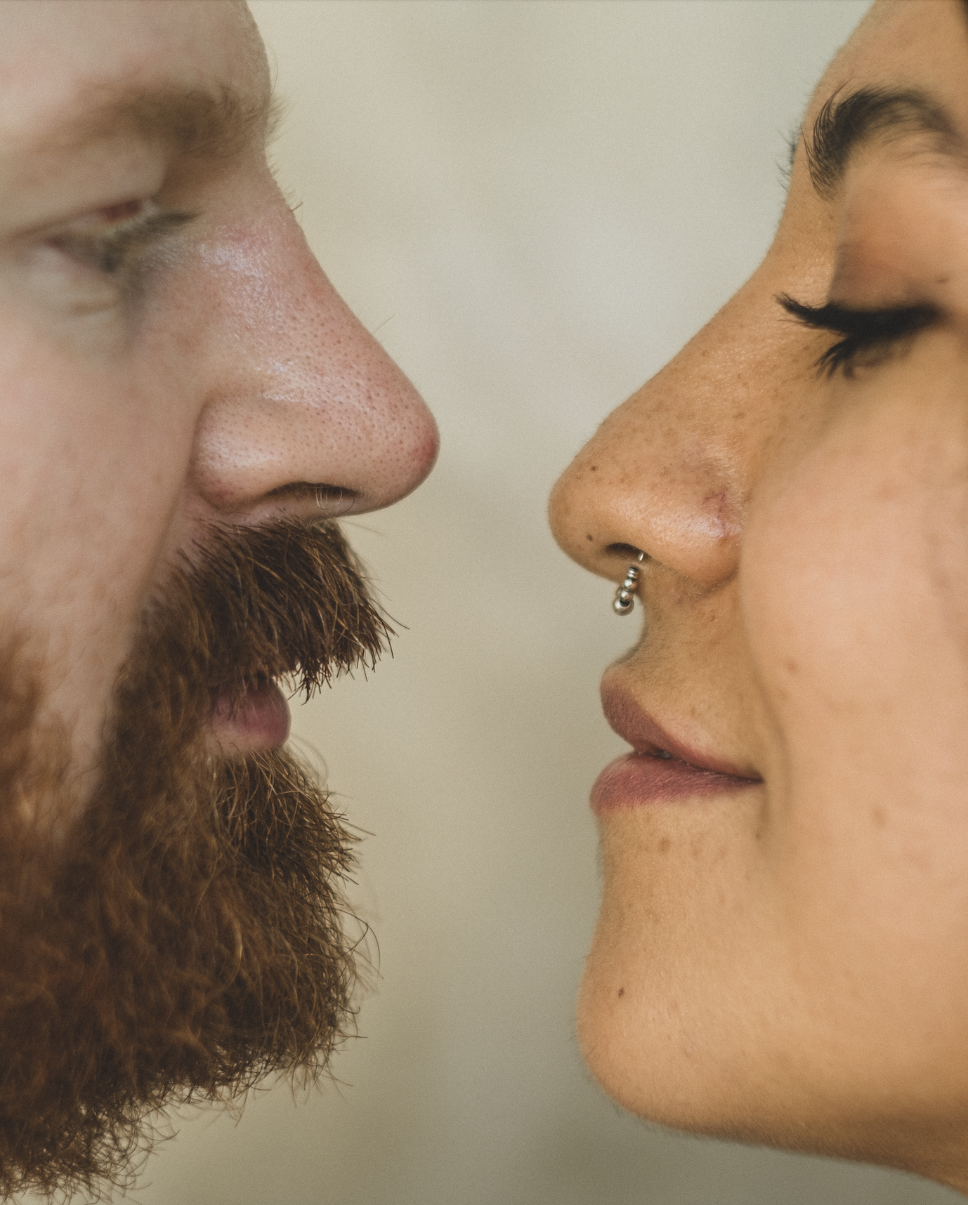 Primer plano de dos personas enfrentadas, nariz con nariz; una tiene barba, la otra un anillo en la nariz; ambas tienen los ojos cerrados, posiblemente a punto de besarse.