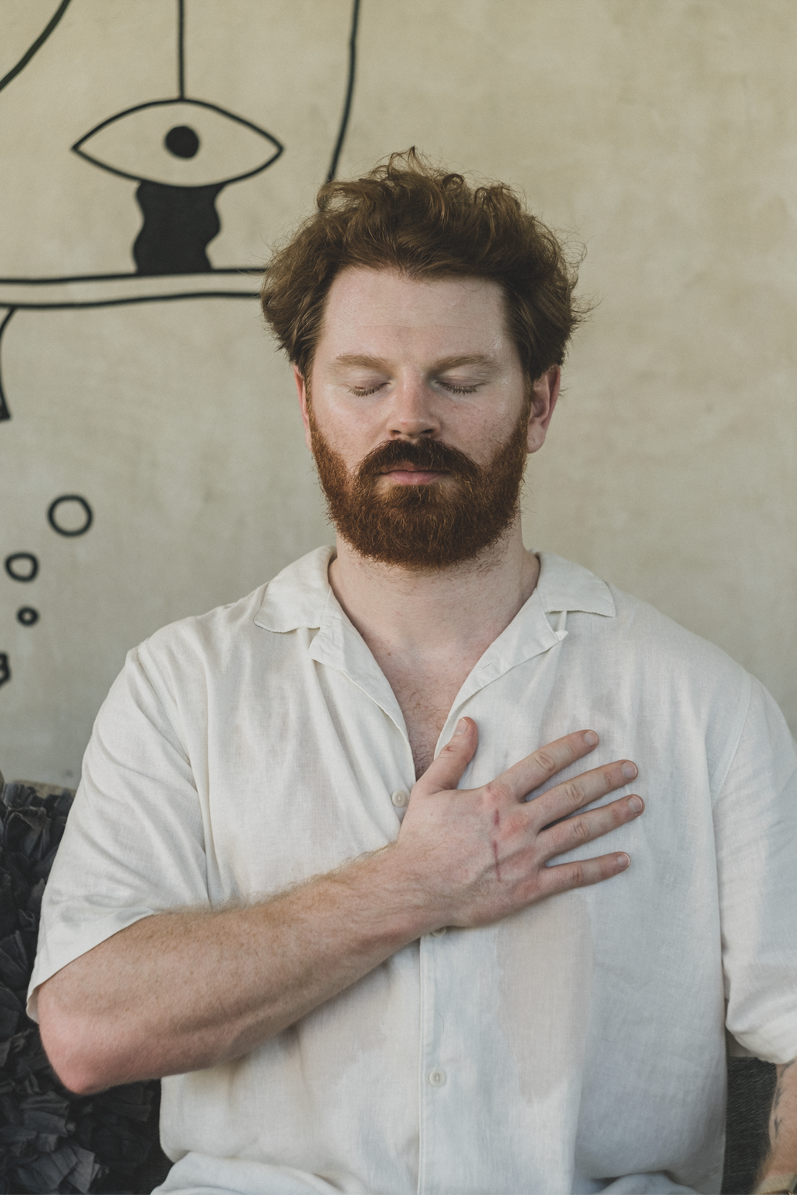 Hombre con los ojos cerrados, la mano en el pecho, meditando. Camisa beige, cabello y barba castaños, frente a un mural.