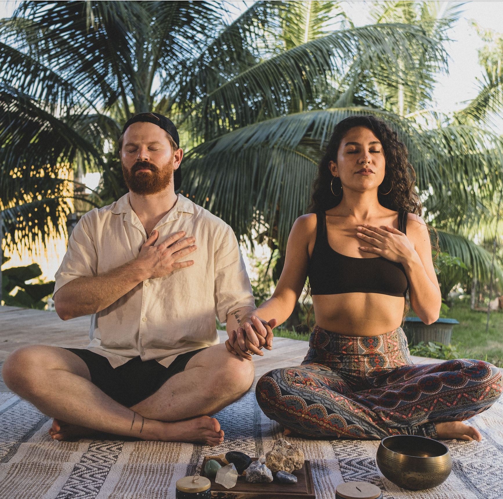 Hombre y mujer meditando, con las manos sobre el corazón, sentados con las piernas cruzadas. Palmeras al fondo.