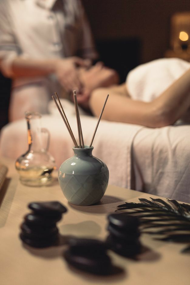 Ambiente de spa: persona recibiendo masaje, barritas de aromaterapia en jarrón, piedras calientes, helecho.