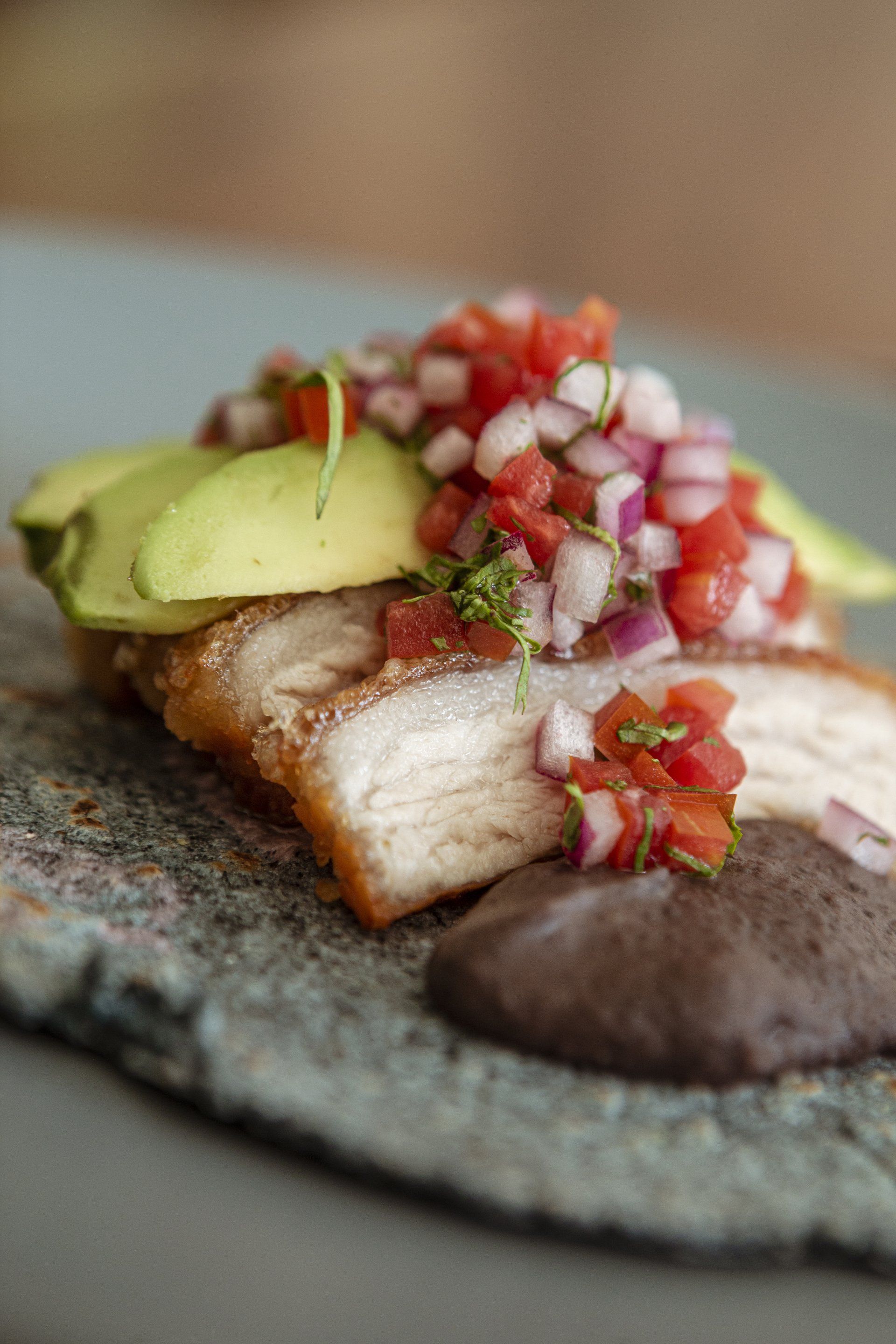 Panceta de cerdo frita cubierta con aguacate y pico de gallo, servida con puré de frijoles negros en un plato rústico.