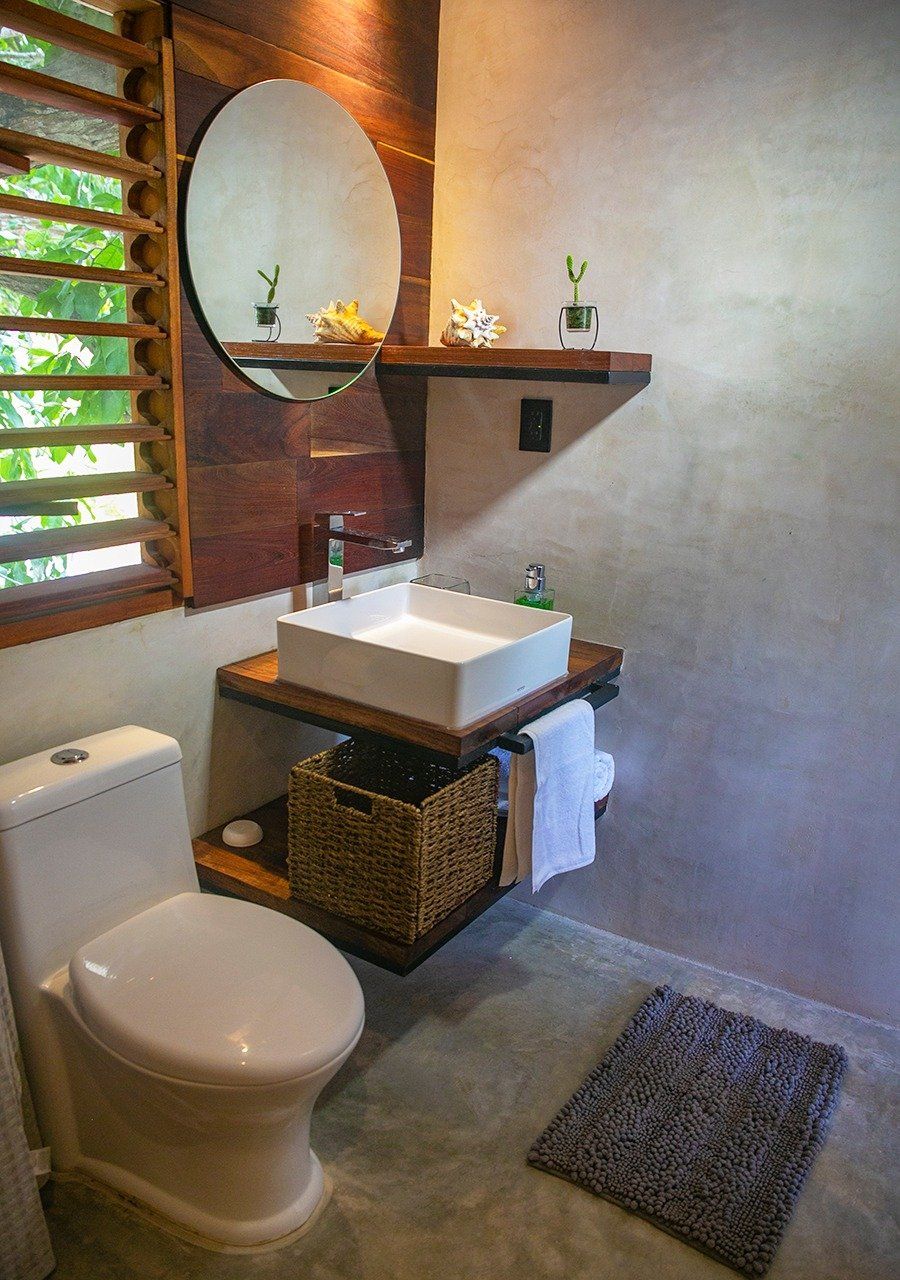 Baño con tocador flotante de madera, lavabo y sanitario blancos; suelo gris; espejo redondo grande.