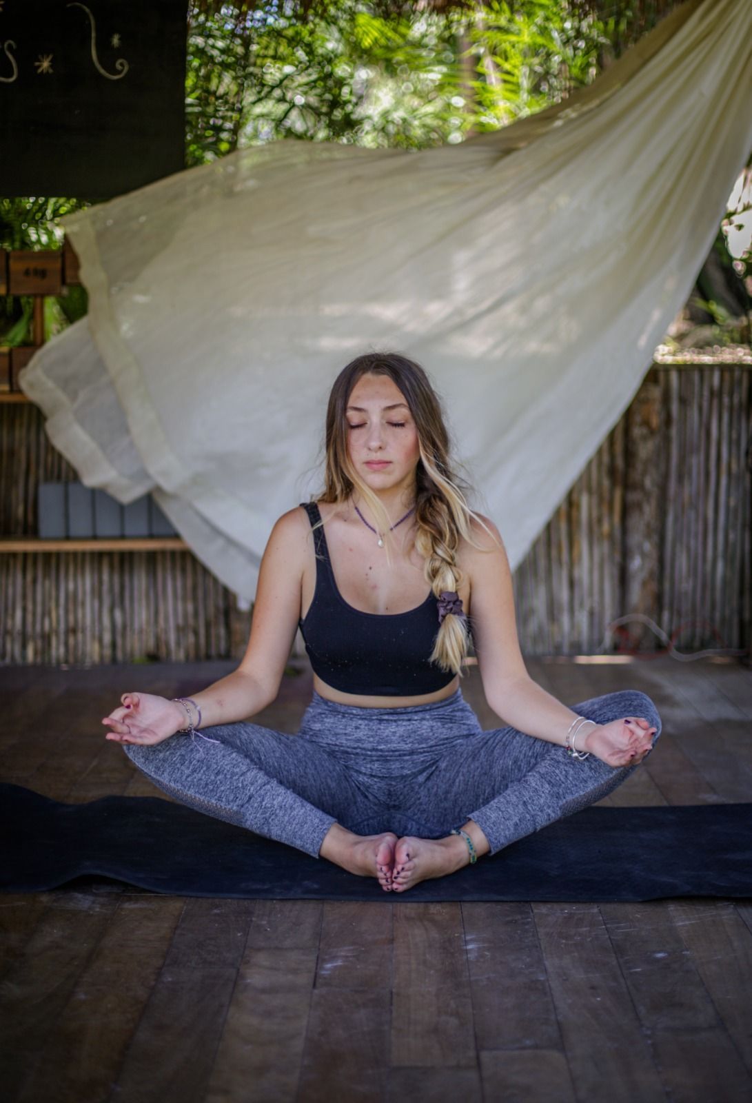 Mujer en postura de yoga, con los ojos cerrados, sobre una esterilla. Lleva sujetador deportivo negro y mallas grises. Al aire libre, suelo de madera y fondo de tela blanca.