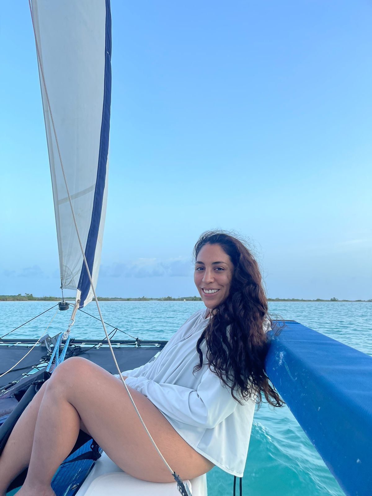 Mujer sonríe en un velero, con el océano al fondo, bajo un cielo azul.
