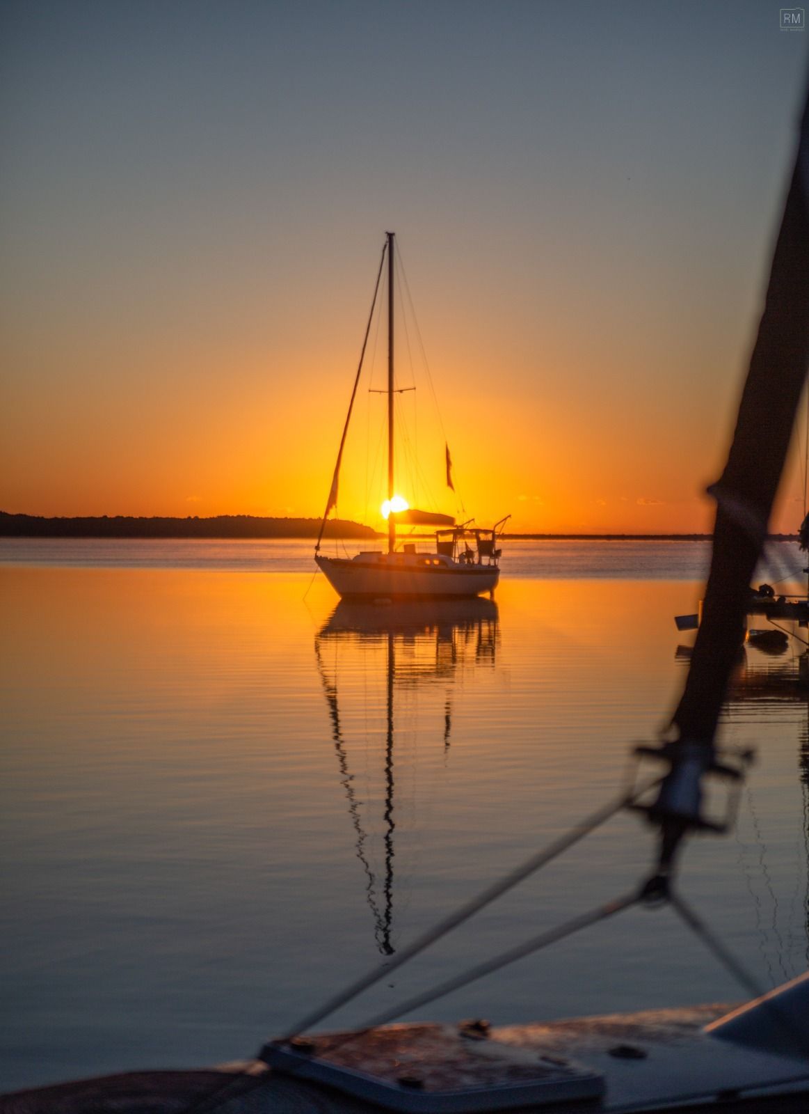 Velero en aguas tranquilas al atardecer, reflejándose en el agua; tonos naranjas y azules.