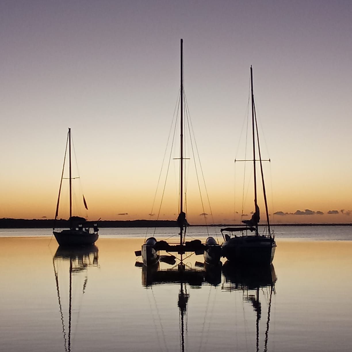 Veleros al anochecer, recortados contra un mar en calma que refleja el cielo naranja y violeta.
