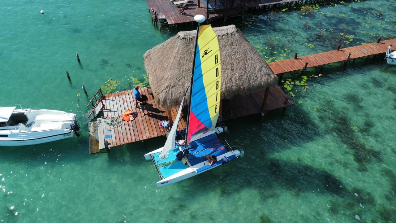 Velero cerca de una estructura con techo de paja en el agua. Vela azul y amarilla brillante, agua turquesa cristalina.