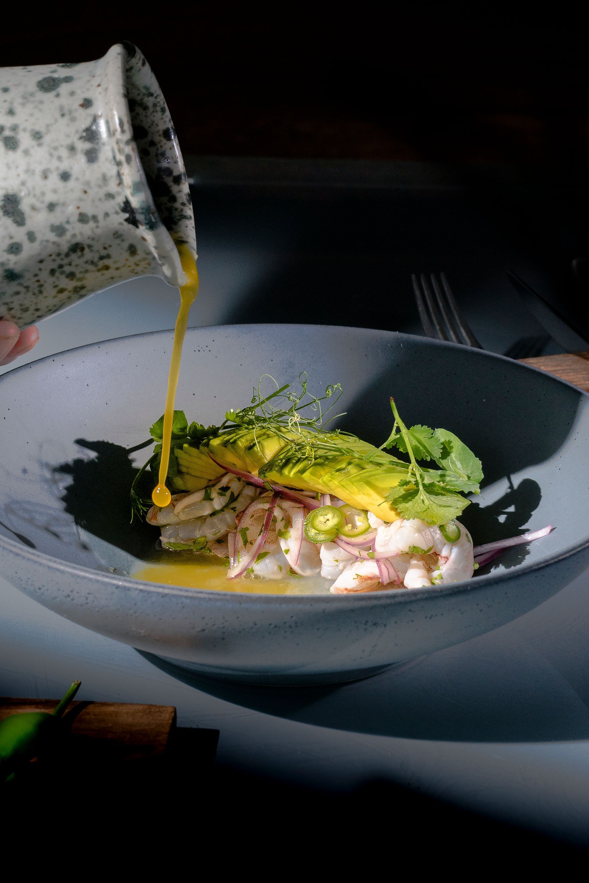 Ceviche rociado con salsa en un tazón azul, adornado con aguacate y verduras. Fondo oscuro.