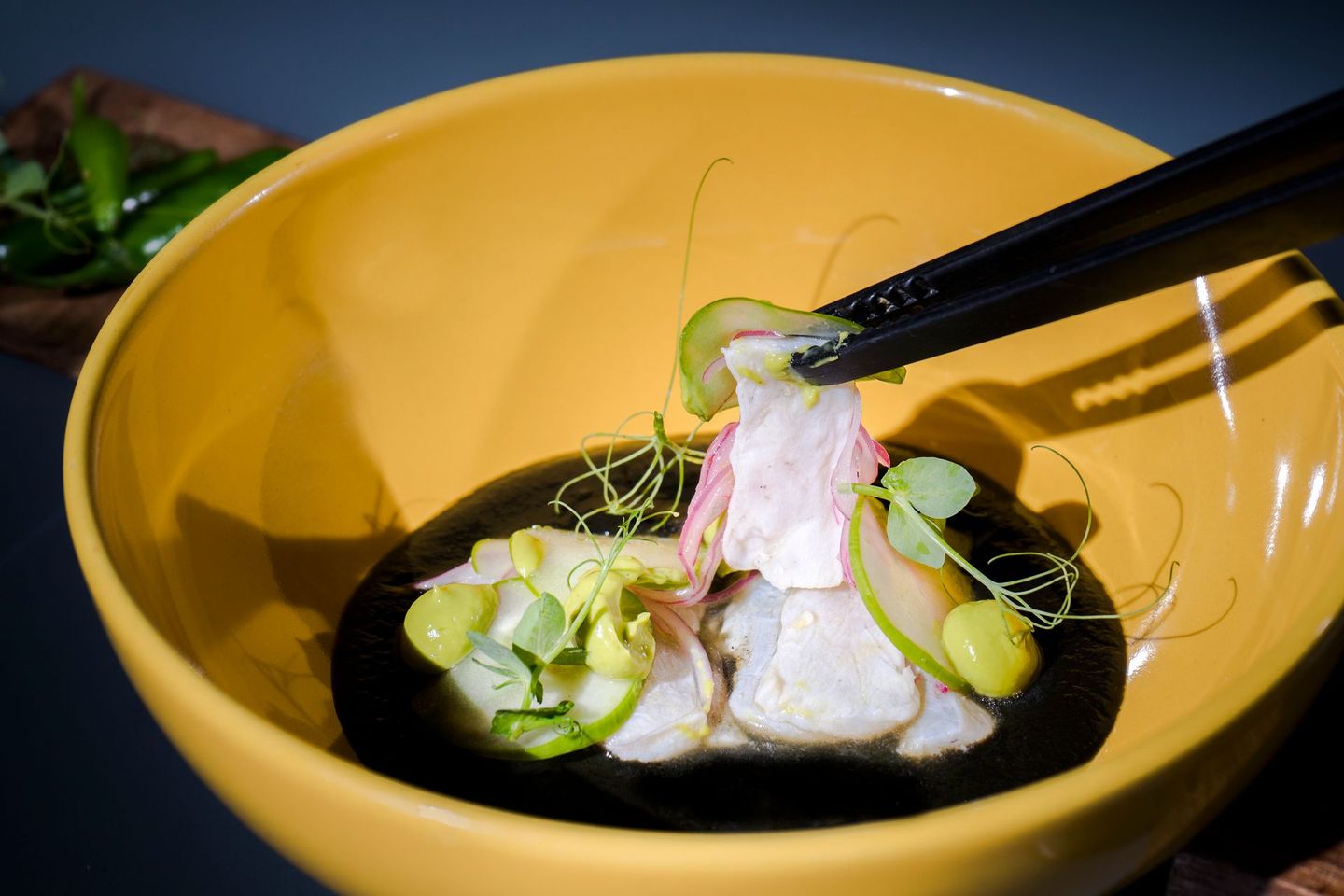 Cuenco amarillo de sashimi, palillos negros levantando un trozo, adornado con verduras y salsa.