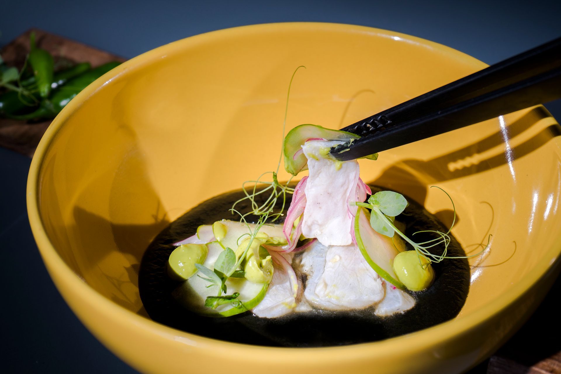 Cuenco amarillo de sashimi, palillos negros levantando un trozo, adornado con verduras y salsa.